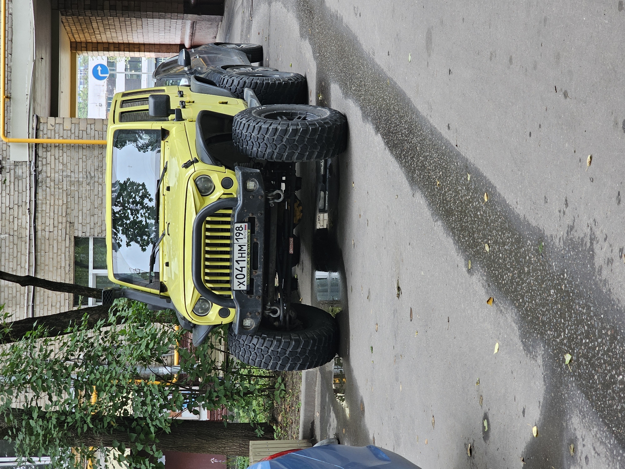 The image shows a bright yellow Jeep Wrangler parked on an asphalt surface, possibly in a courtyard or under an overpass, with a wet sheen indicating recent rain. The Jeep is modified with large, aggressive off-road tires and a snorkel air intake. A dark-colored sedan is parked to the right of the Jeep. In the background, a brick building with a yellow pipe running along its facade is visible. Trees with green foliage are also present on the left side of the image. A blue and white traffic sign with a right arrow is visible further in the background, suggesting an urban environment. The license plate of the Jeep reads "X041HM 198 RUS". The scene is static with no people present.