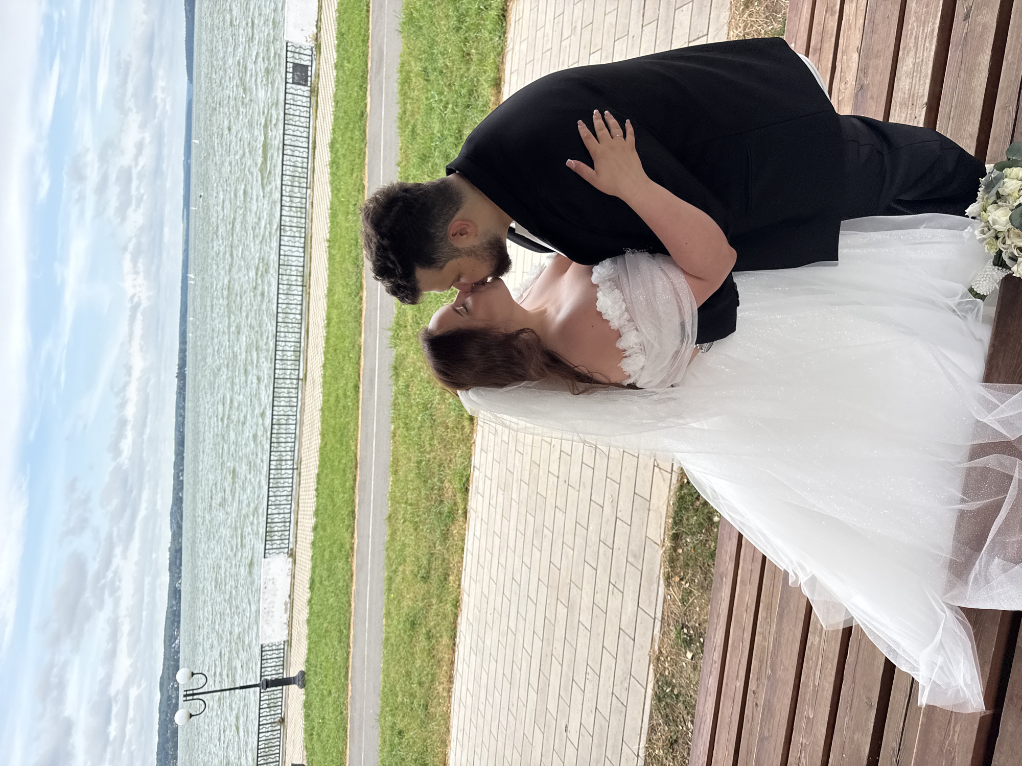 A couple is kissing on a wooden bench overlooking a large body of water in Votkinsk, Russia. The woman is wearing a white wedding dress and veil, and the man is in a black suit. The water is calm with a few sailboats visible in the distance. The shore is lined with a paved walkway and a metal fence. The sky is cloudy.