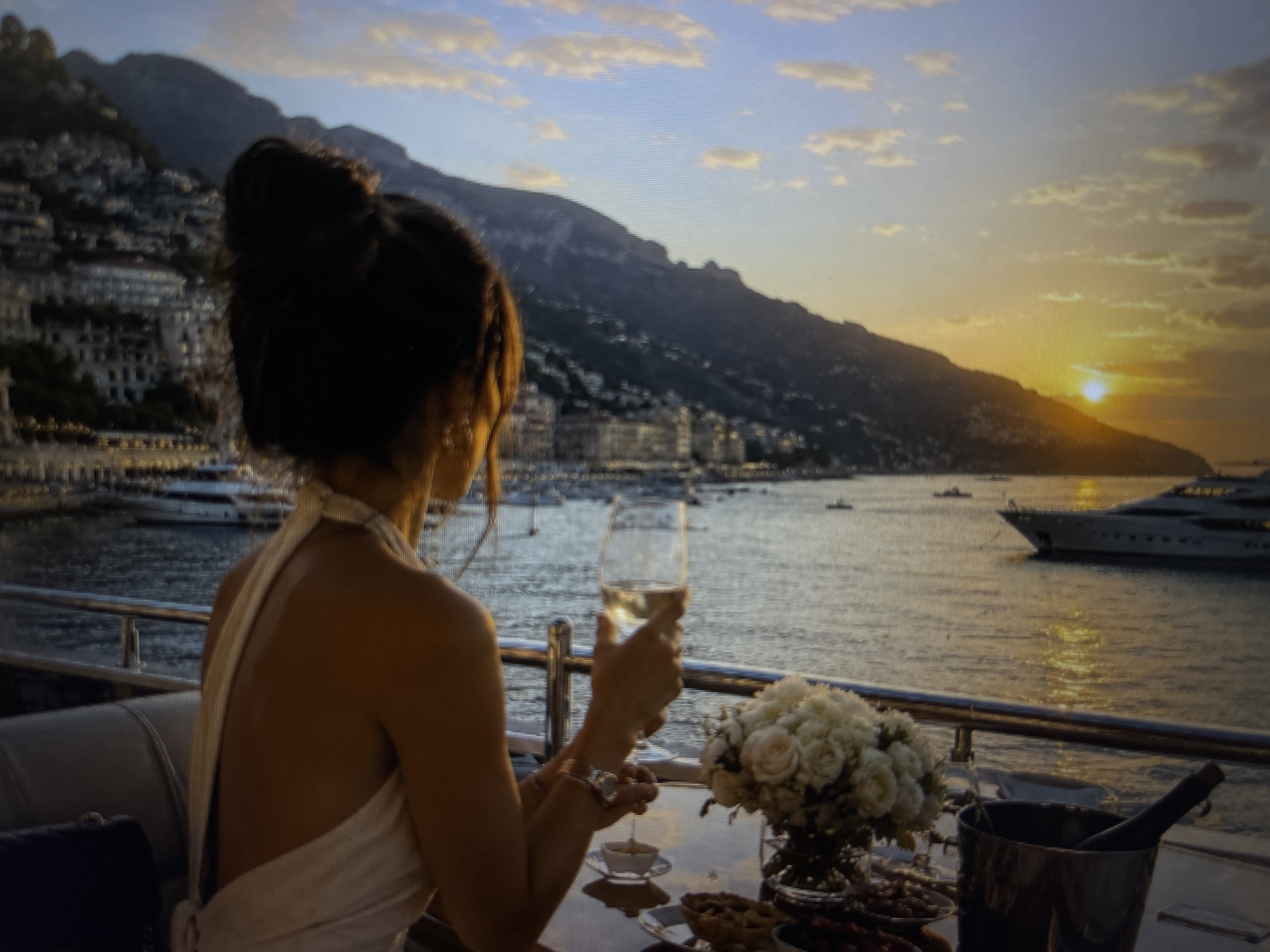 A woman with dark hair in a high bun is sitting on a yacht, facing away from the viewer and holding a glass of white wine. She is wearing a white halter-top dress. In front of her is a table with a vase of white flowers, a small dish, and a silver ice bucket with a bottle inside. The yacht is on the water, with a coastal city and mountain range in the background. The sun is setting, creating a golden glow. The location of this scene is not in Sofia, Bulgaria, but appears to be a coastal location in Italy, possibly Positano.