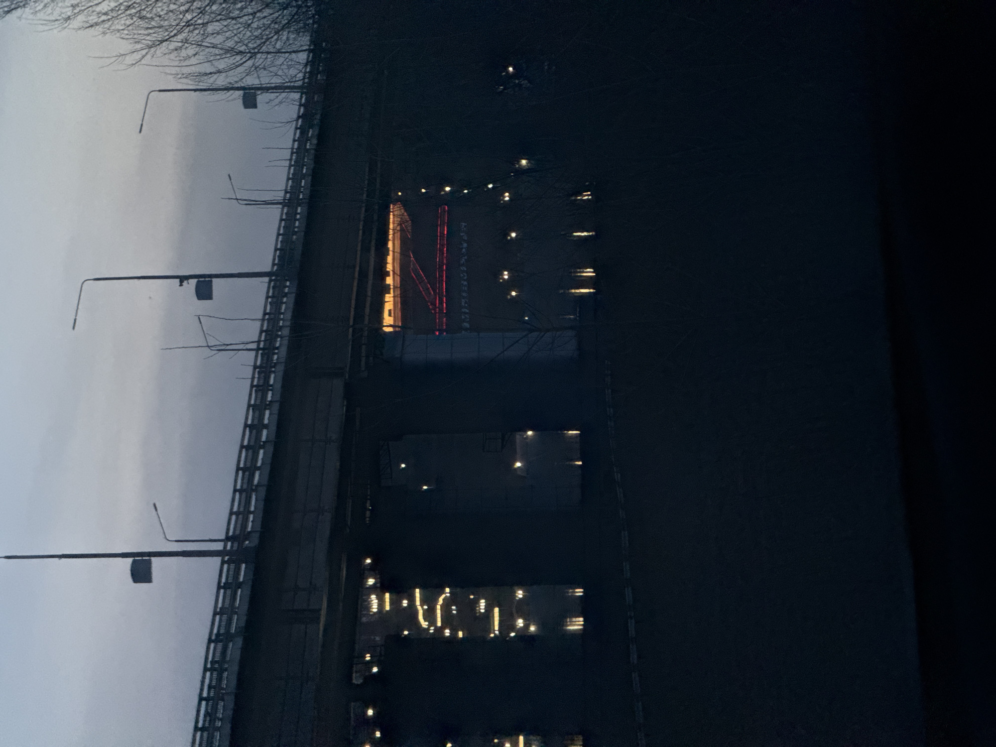 This image, rotated 90 degrees counter-clockwise, depicts an outdoor scene at dusk or night, viewed from an elevated vantage point, likely a bridge or overpass, looking across a body of water towards a distant shore.
In the upper portion of the frame (when upright), bare tree branches are silhouetted against a cloudy, grey sky. Below the trees, a dark bridge structure with railings and multiple lampposts extends across the scene. The lampposts are dark, but some have small, box-like structures attached to them.
The lower portion of the image is dominated by a dark body of water, possibly a river or bay, which reflects the lights from the opposite bank. On the distant shore, several buildings and structures are illuminated. A prominent, tall building on the left glows brightly with warm yellow light. Adjacent to it, a striking red, stylized letter, resembling an 'N', is brightly lit and stands out. Further along the shore to the right, more buildings are visible, their numerous lighted windows casting vertical streaks of light onto the dark water. Scattered smaller lights also dot the water's surface, likely reflections of distant sources.
The time of day is clearly evening or night, indicated by the dark sky and the reliance on artificial lighting. The weather appears overcast, with no stars visible. There are no people visible, and no specific activities are discernible, creating a quiet, somewhat serene urban or industrial nightscape. No other explicit text is visible beyond the stylized red 'N'.