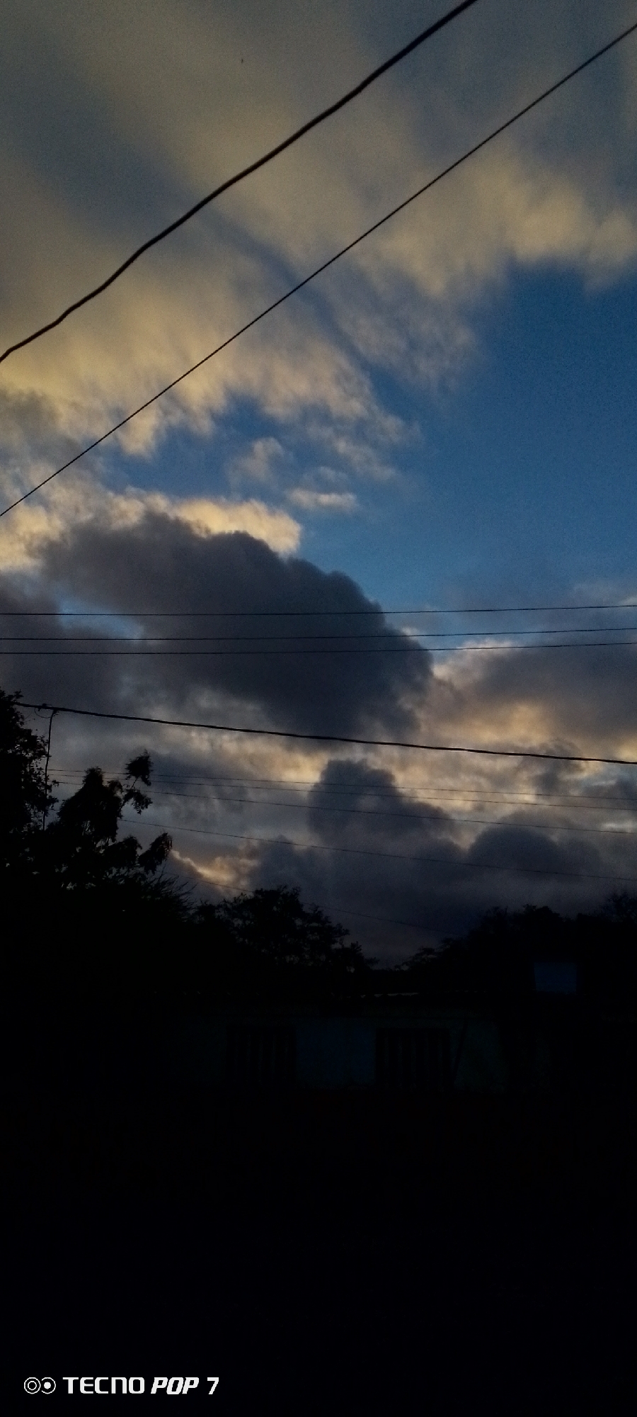 The image shows a twilight sky with scattered clouds, illuminated by the setting sun. Two power lines run diagonally across the frame. Silhouetted against the sky are trees and a building. The text "TECNO POP 7" is visible in the lower left corner, indicating the device used to capture the image. The scene is likely located in Altagracia, Venezuela, given the reference.
