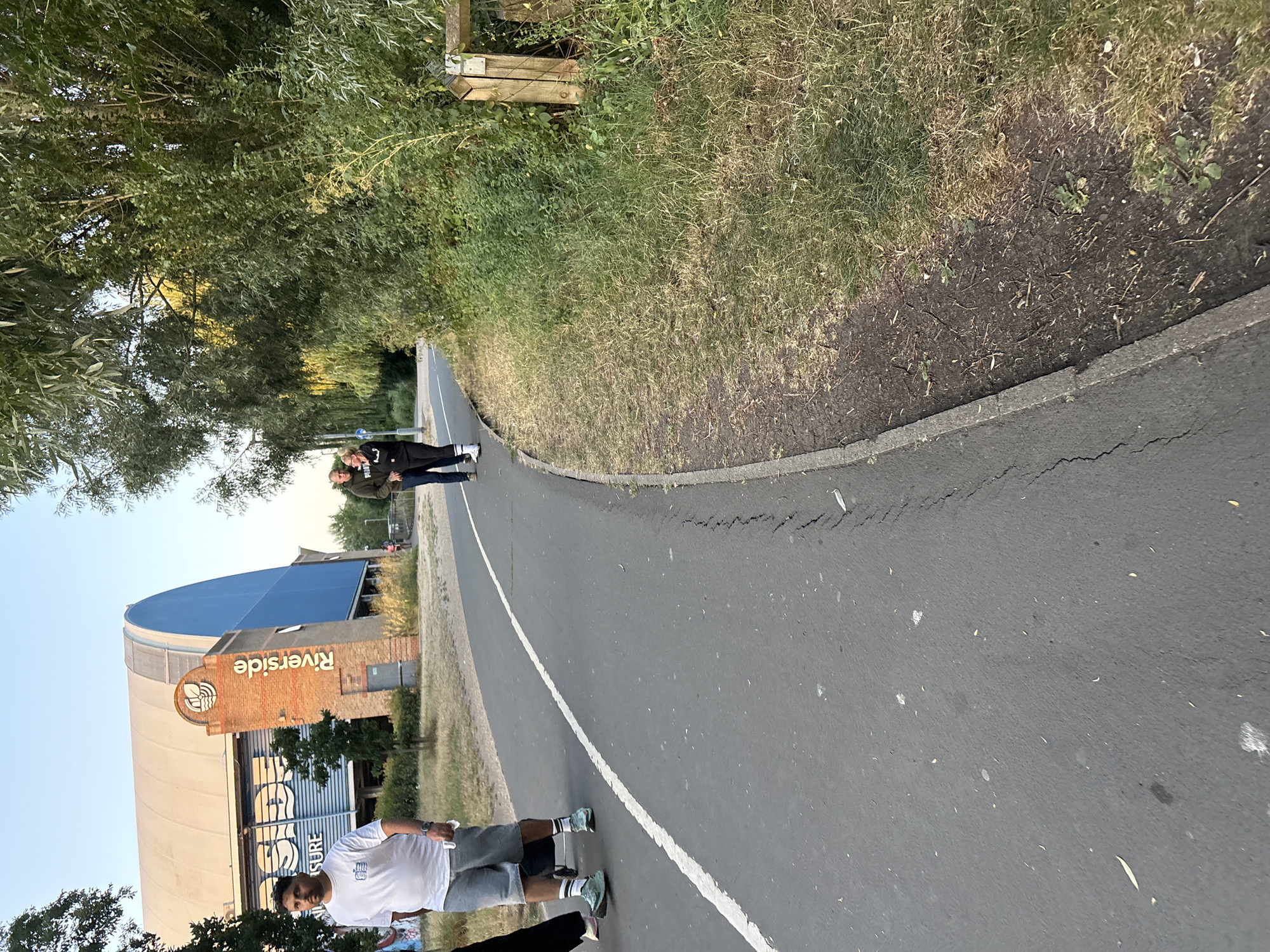The image shows a paved pathway with a white line marking its center, curving to the right. On the left side of the pathway, a man wearing a white t-shirt and grey shorts is walking, pushing a black wheeled bag. Further down the pathway, a couple is walking together, the man in a dark hoodie and the woman in a dark top and black pants.
In the background on the left, a large building with a curved, light-colored roof and a blue section is visible. A brick pillar with the word "Riverside" in vertical lettering and a circular logo with a leaf design is attached to the building. The building appears to be the Riverside Leisure Centre in Chelmsford, United Kingdom.
To the right of the pathway, there is a grassy area with dry grass and some green vegetation. A wooden post and wire fence is also visible in this area, near a gate-like structure. Trees and dense foliage line the far right of the scene, with some sunlight filtering through the leaves. The sky is clear and light blue, indicating daytime.