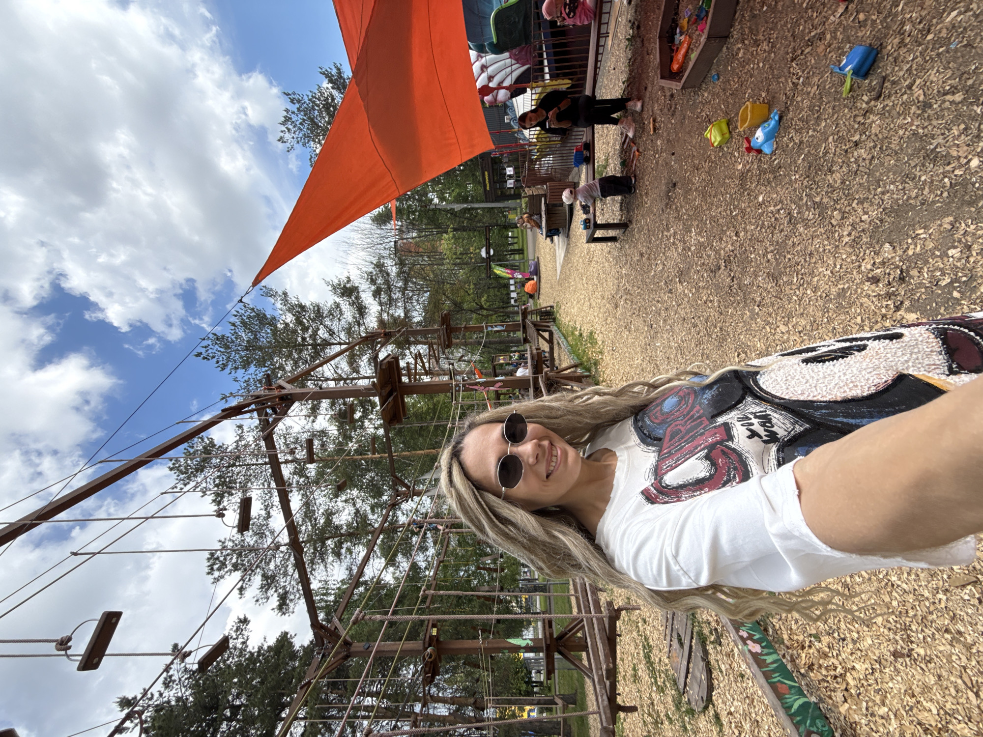 A selfie-style image captures a woman smiling in an outdoor adventure park setting in Шебунино, Russia, on a bright day. The woman, with long blonde hair and wearing sunglasses and a white t-shirt featuring a stylized graphic possibly related to Mickey Mouse (with visible elements resembling "Mickey" and "POP"), is positioned in the foreground, facing the camera.
The background reveals a recreational area with various features. A prominent orange shade sail extends over part of the scene, providing cover. Below it, several individuals, including adults and children, are visible. One adult, dressed in dark clothing, stands near a sandpit filled with toys like buckets and spades. A child is also seen near a small table.
To the left and extending into the middle ground is a large wooden structure, appearing to be an aerial ropes course or climbing frame, with ropes, wooden steps, and platforms. Trees are scattered throughout the background, indicating a natural environment. The ground is covered with wood chips, typical of playgrounds and adventure parks. The sky is blue with scattered white clouds, suggesting pleasant weather conditions suitable for outdoor activities. The overall mood appears joyful and relaxed.