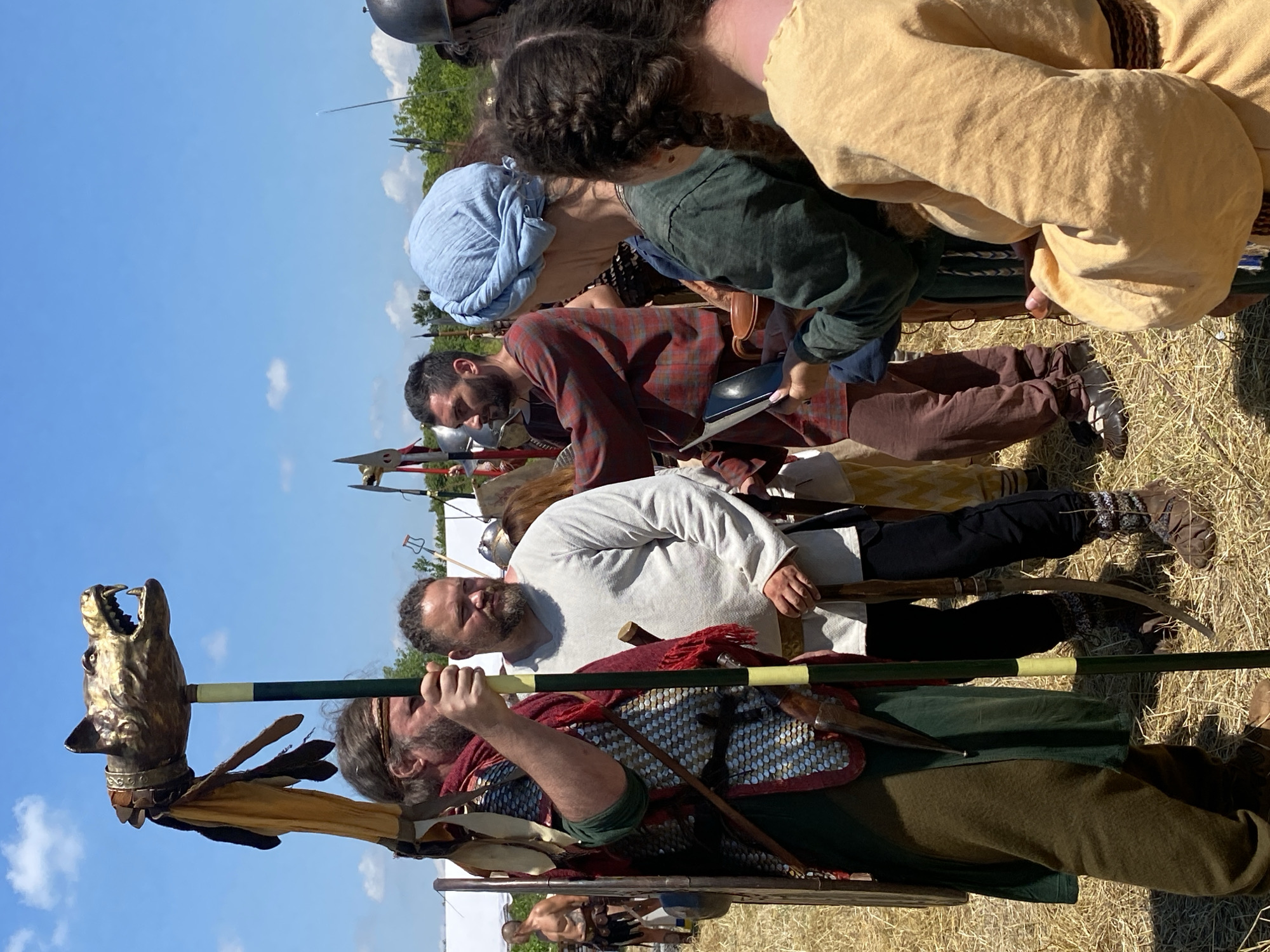 The image depicts a historical reenactment event taking place outdoors under a clear blue sky with sparse white clouds. The location is Gigen, Bulgaria.
In the foreground, slightly left of center, a man with a reddish-brown beard and long hair, wearing a scale armor tunic over a green undergarment and a red scarf, holds a tall staff vertically. The staff is topped with an ornate golden wolf's head, snarling with bared teeth, and adorned with dark feathers and yellow fabric strips. A round, plain wooden shield is partially visible on his left arm. He wears brown trousers and brown shoes.
To his right, a man with a dark beard and dreadlocks, wearing a plain white long-sleeved tunic and dark trousers, stands with a wooden staff or tool, possibly a digging implement, in his right hand. He wears patterned socks and brown, soft-soled shoes.
Further right, a man with short dark hair and a beard, dressed in a red plaid long-sleeved tunic and brown trousers, stands facing partially right. He appears to be interacting with another individual or an object not fully visible. He wears dark shoes.
In front of him, facing away from the camera, is a person wearing a light blue fabric head covering, a green tunic, and possibly blue undergarments. This individual is looking down, holding a small dark, rectangular object (possibly a book or tablet) in one hand and a small, reddish-brown clay pot or cup in the other.
To the immediate right of this figure, another person with dark braided hair (pigtails) and a light yellow/beige long-sleeved tunic stands, also facing away and looking down.
In the far background to the left, a shirtless man with a beard and dark trousers or kilt, possibly adorned with animal skins, stands near a white tent. In the mid-ground, various other figures are partially visible, including one on the far right wearing a helmet, and several upright poles, possibly spears or tent supports. The ground is covered with dry, yellowish straw or cut hay. The overall scene suggests a vibrant historical gathering or encampment during daylight hours.