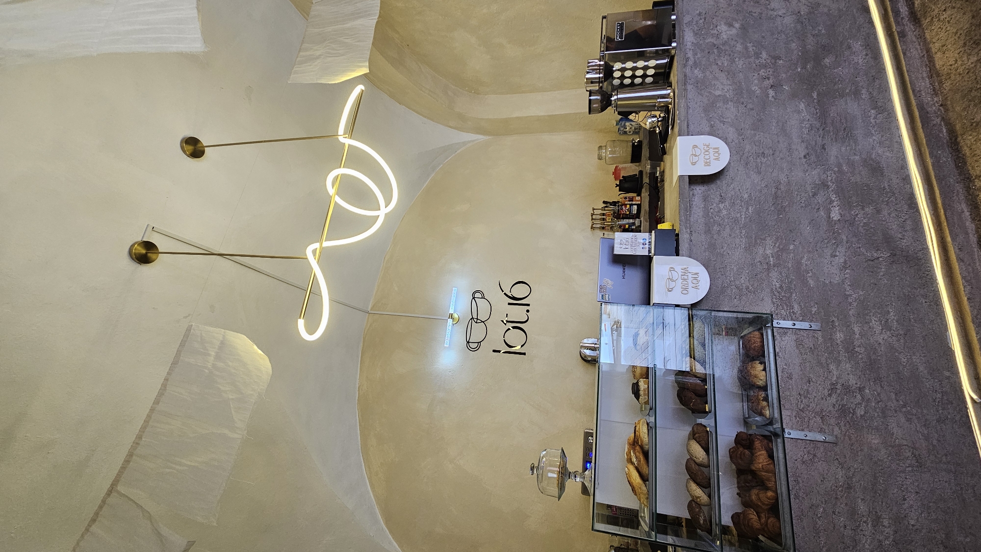 This media file captures the interior of a modern coffee shop or bakery, likely located in Puebla, Mexico, given the Spanish text. The perspective is rotated 90 degrees clockwise, presenting the wall as the base of the image.
The main subjects include an arched, light-colored ceiling with textured details and modern, abstract lighting fixtures featuring glowing white tubes and brass elements. On a textured, light beige wall, a prominent logo "lot.16" is displayed, accompanied by an icon of two coffee cups, some of which is also rendered as a neon sign.
Below this, a service counter extends across the image. On the counter, a professional "ROCKET" brand coffee machine is visible, along with various bottles and jars for ingredients. In the foreground, a multi-tiered display case showcases a variety of baked goods, including croissants and different types of bread.
Two white, coffee cup-shaped signs are placed on the dark, textured counter. One reads "ORDENA AQUÍ" (Order Here) and the other "RECOGE AQUÍ" (Pick Up Here), indicating specific service points. A device with "HUAWEI" branding is also partially visible behind the counter.
No people are present, suggesting the image might have been taken during opening hours before customers arrived or after closing. The overall impression is one of a clean, stylish, and inviting commercial space. The lighting from the fixtures and neon sign contributes to a warm, welcoming atmosphere.
