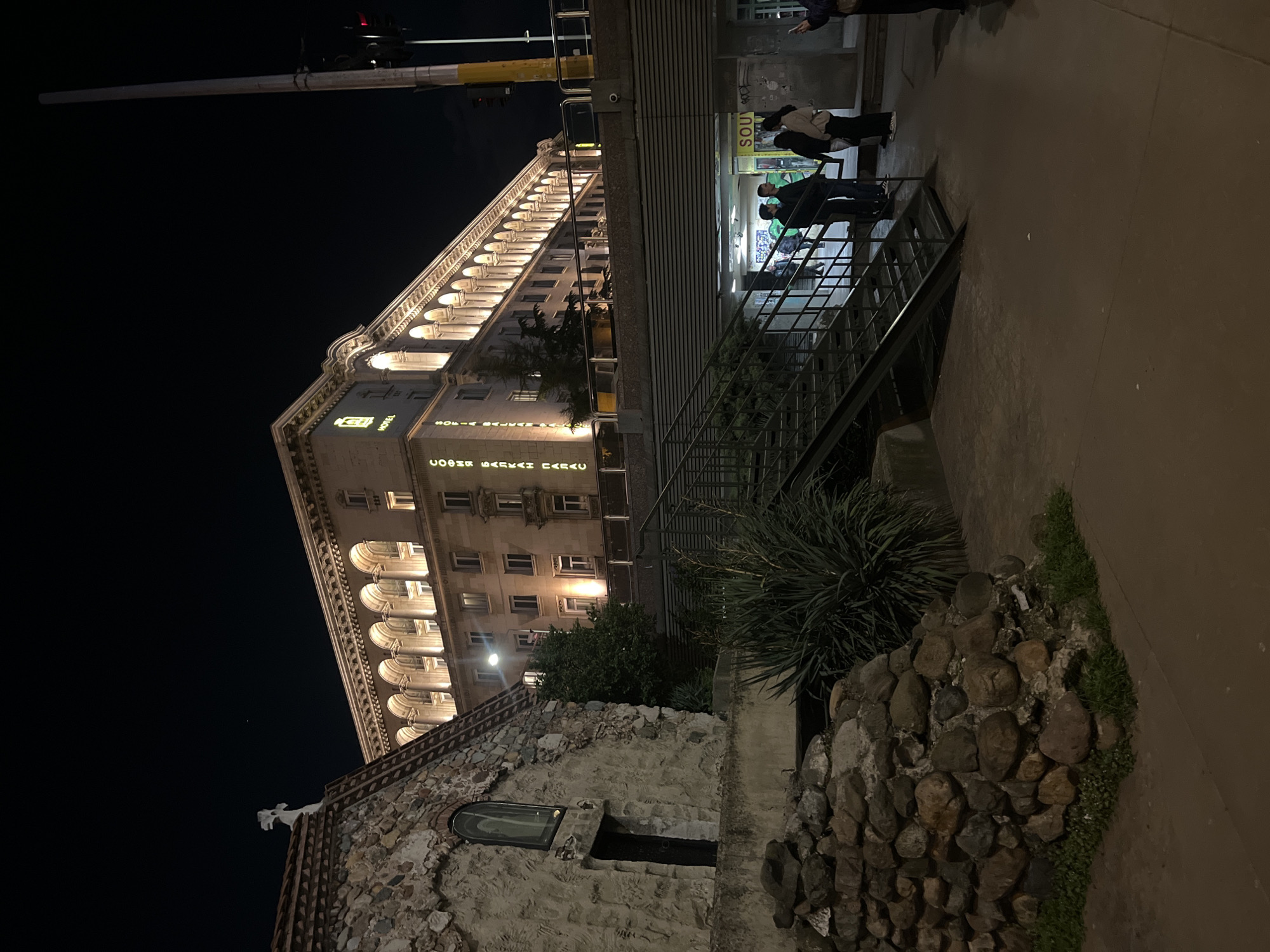 This nighttime image depicts an urban street scene in Sofia, Bulgaria, characterized by a blend of grand architecture and historical elements under artificial illumination.
**Subjects**: The main subject is a large, multi-story building, likely a hotel, dominating the background. Its ornate facade features numerous arched windows and is brightly lit, highlighting architectural details. In the foreground, to the left, stands a smaller, rustic stone-built structure, possibly an ancient ruin or monument, featuring a small arched window and a cross on its roof. A gargoyle-like figure is visible near the top edge of this stone structure. Several individuals are present on a raised concrete walkway or platform to the right, appearing to be standing or casually walking. Greenery, including bushes and patches of grass, is visible around the stone structures and near a rock-built planter in the immediate foreground. Street lights or traffic signals are present in the upper left.
**Activity or event**: The activity is limited to people standing or walking on the elevated walkway. No specific event is discernible.
**Notable details**: The time of day is clearly night, with the sky completely dark and all illumination coming from artificial light sources, casting a warm glow on the buildings. The image conveys an urban setting with a mix of historical architecture and modern infrastructure like the metal staircase and walkway. The location context is Sofia, Bulgaria.
**Visible text**: On the large illuminated building, the text "SOFIA BALKAN PALACE" and "HOTEL" is clearly visible. Below the elevated platform, an entrance to a shop or underground area shows partially legible text, "SOUT".