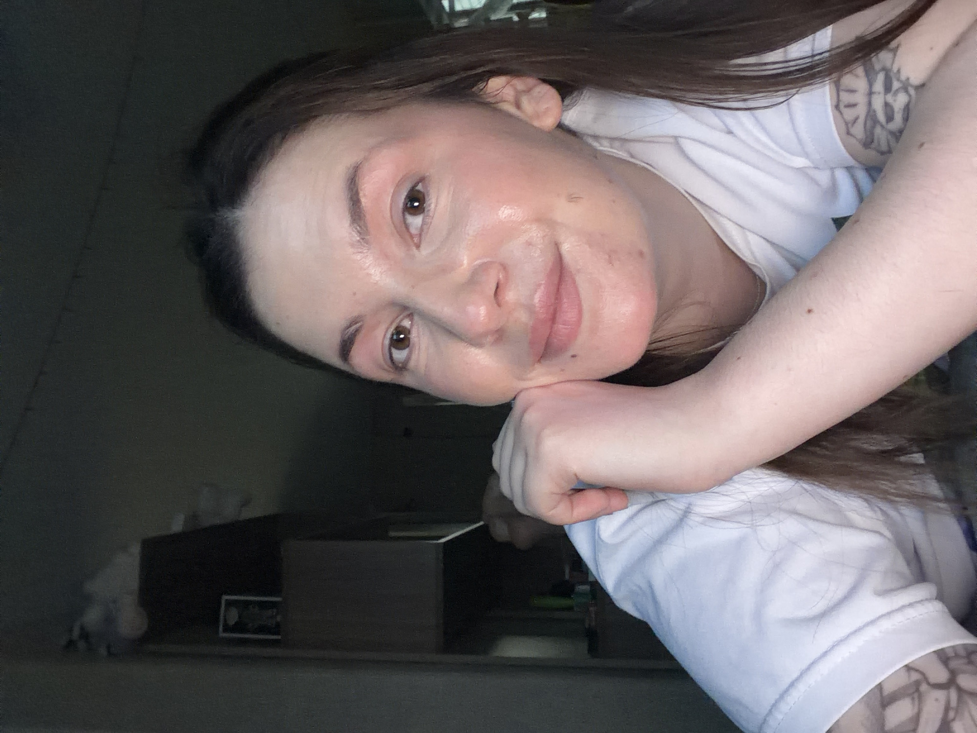 A person is posing for a selfie. The person has brown hair, brown eyes, and is wearing a white t-shirt. The person has a tattoo on their arm. The background is dark and indistinct, but appears to be a room with furniture. There is no indication that the location is Chaikovsky, Russia.