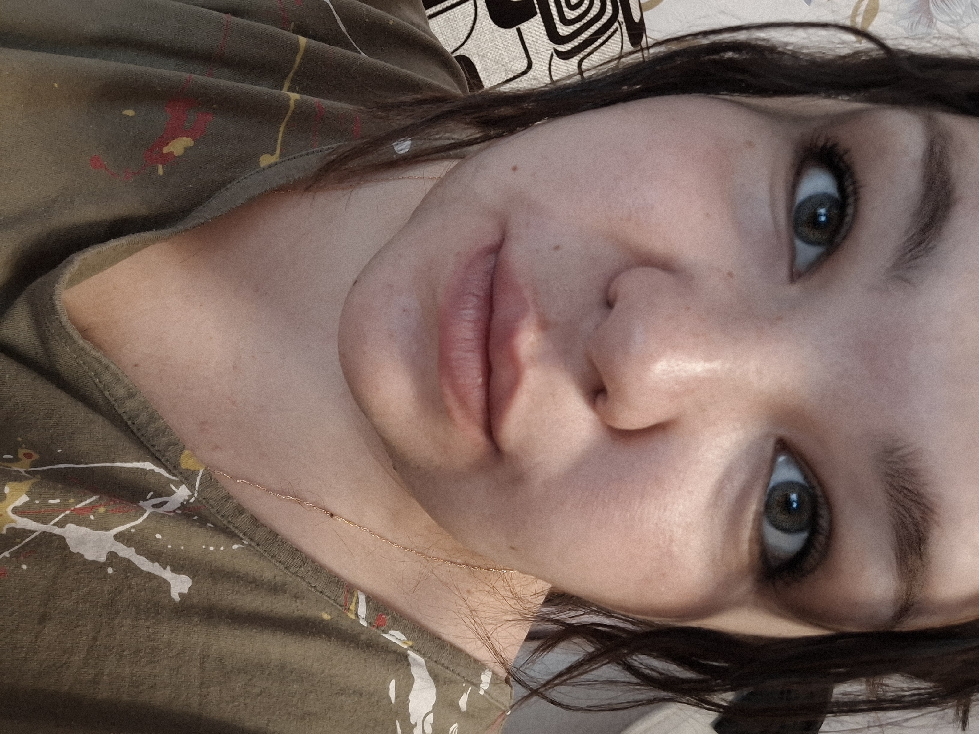 A close-up selfie captures a woman's face. Her eyes are large and framed by dark eyelashes and eyebrows. Her lips are full and unpainted. A thin gold chain necklace is visible against her skin. She wears a green t-shirt with paint splatters. The background shows a blurred, patterned headrest. There is no indication that this image was taken in the city of Марковский, Russia.