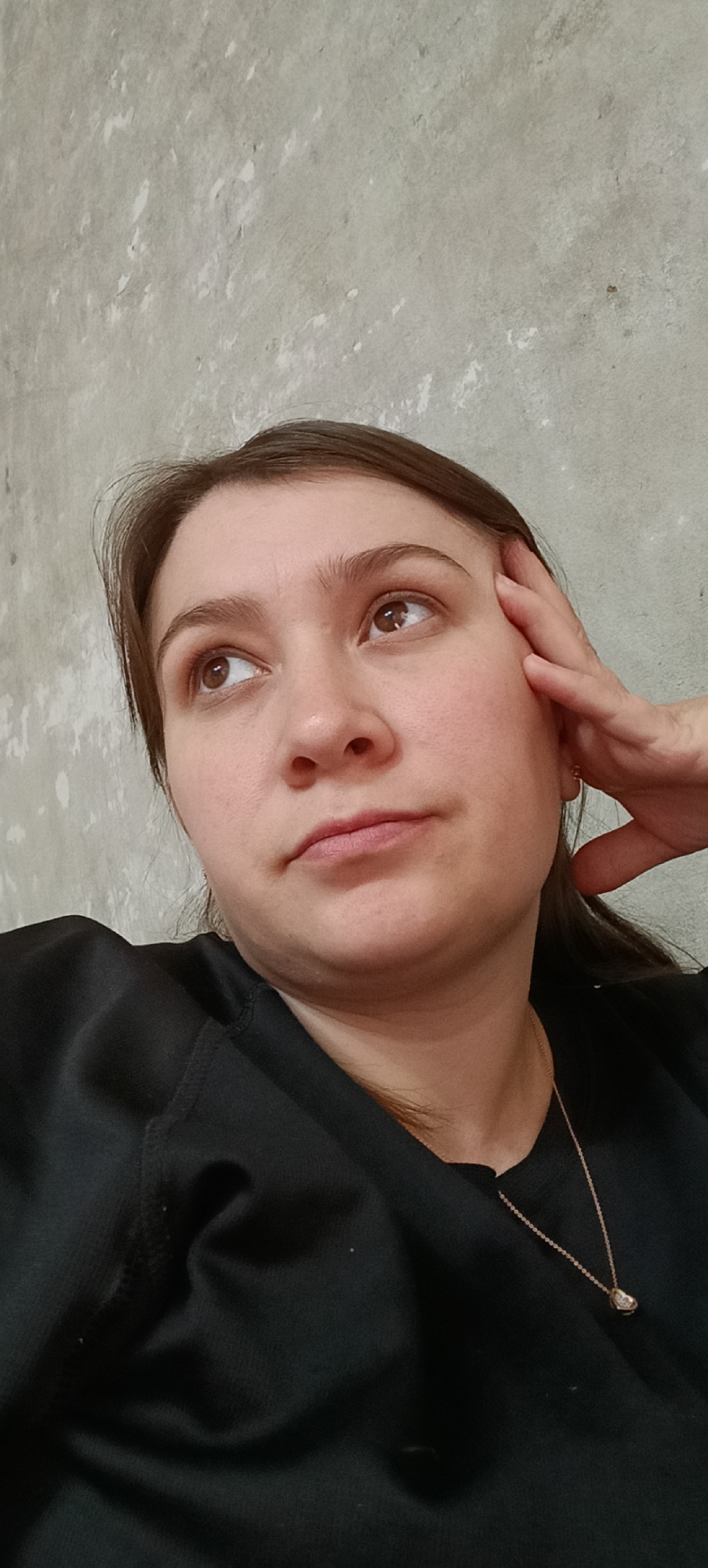 A woman with brown hair is resting her right hand on her forehead, looking up and to her left. She is wearing a black top and a delicate gold necklace with a small pendant. The background is a textured, light gray wall. The image was taken indoors.