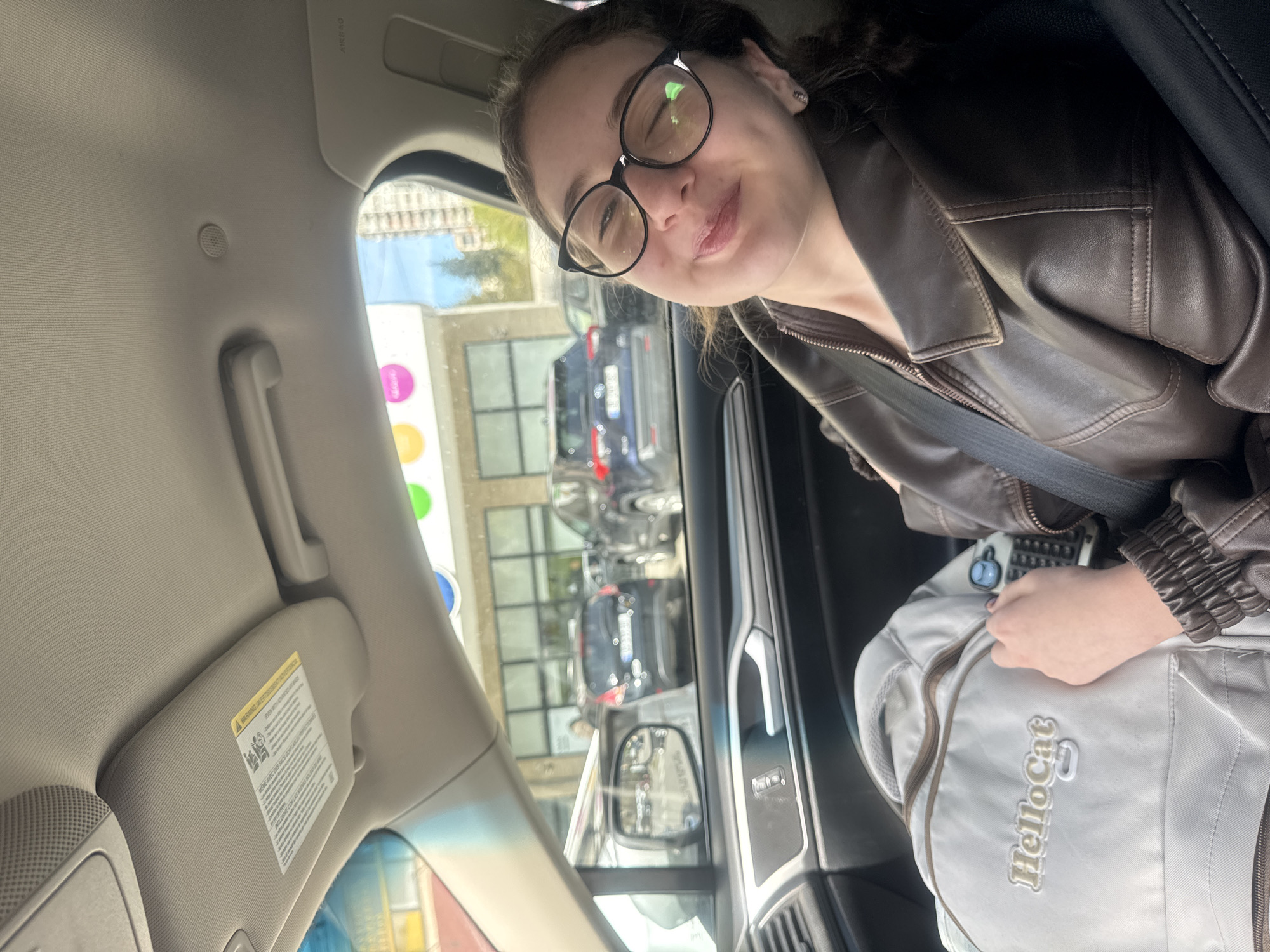 The image captures a young woman, appearing to be in a car, likely in Tbilisi, Georgia, during the daytime under clear skies. She is positioned as a passenger, smiling softly towards the camera. She wears black-framed glasses, through which her eyes are visible, and has dark hair, part of which is pulled back. She is dressed in a brown leather jacket and is wearing a seatbelt across her chest.
Her left hand rests on a light gray backpack that is visible on her lap or beside her. The backpack has the text "HelloCat" embroidered in gold lettering on its front.
The car's interior is visible, showing a light-colored headliner, a grab handle, and a sun visor with a yellow warning label. The "AIRBAG" label is visible on the A-pillar. Through the driver's side window, an urban street scene is observed, with several parked cars, buildings, and a clear blue sky. A side mirror and a portion of the rearview mirror reflect parts of the exterior. Outside, there's a building with some colorful circular signage (purple, green, blue, yellow) that might belong to a business. The overall impression is one of a casual moment, possibly waiting or traveling in a car.