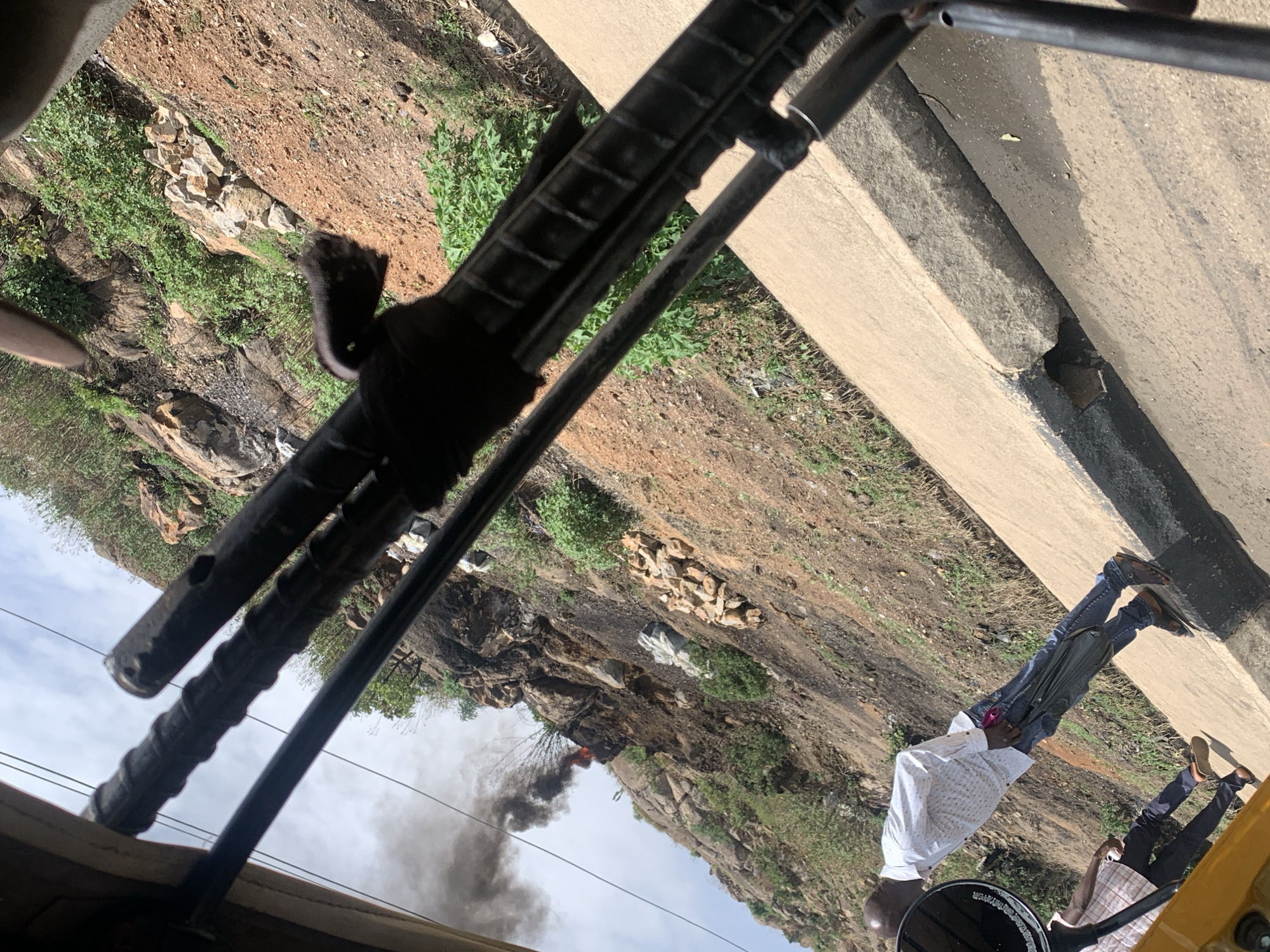 The image, likely taken from within a vehicle, shows a scene near Gura Top, Nigeria. A roadside curb and adjacent vegetation are visible. In the distance, a hillside shows evidence of a fire with smoke billowing upwards. Two individuals are visible on the right side of the frame, near the roadside. The perspective is angled. Elements of the vehicle's interior are in the foreground.