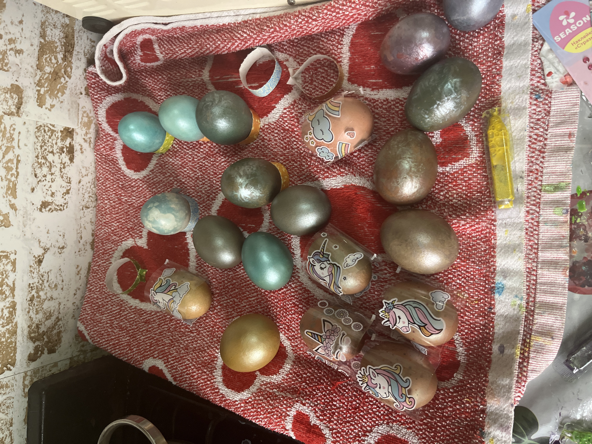 The image depicts a collection of Easter eggs, some of which are decorated with unicorn stickers, arranged on a red and white patterned towel. Other eggs are painted in metallic colors such as silver, gold, and teal. There is also a tube of yellow paint. A "SEASON" sticker sheet is visible on the right side. The scene appears to be indoors, possibly in a home. The location is Oryol-Izumrud, Russia.