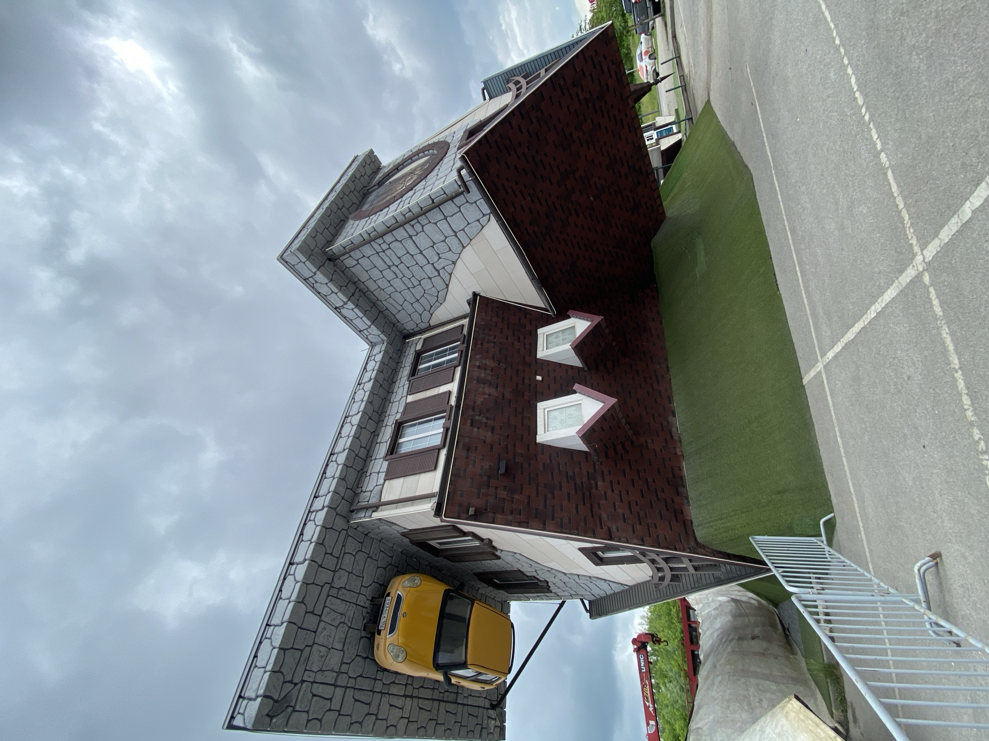 This is an external, low-angle, wide shot of an upside-down house attraction located in Ufa, Russia. The house is a tourist attraction, designed to look like it is built upside down, with the roof and chimney on the ground and the foundation in the air. A yellow car is parked on what appears to be the roof. The house has a stone-like facade on the top, and brickwork on the lower part. The sky is overcast and cloudy. In the foreground, there is a grey metal fence and a paved area. In the background, there are trees, a red crane, and some vehicles parked. The overall impression is one of playful architectural illusion.