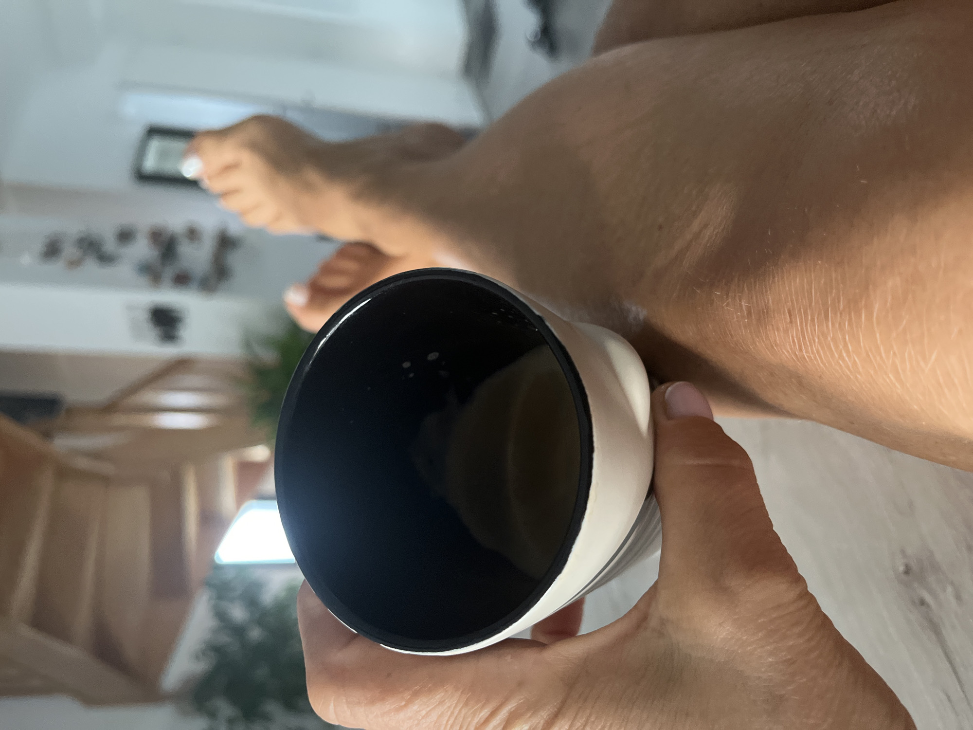 In the city of Chambœuf, France, a close-up shot captures a person's hand holding a white and black mug filled with a light-colored liquid, possibly coffee or tea. The mug is positioned in the foreground, with the person's legs visible in the background, crossed and resting. The skin of the legs appears tanned, with a faint shimmer suggesting moisturizer or lotion. A staircase with wooden railings is visible in the blurry background, hinting at an indoor setting. The overall lighting suggests a bright, possibly daytime scene.