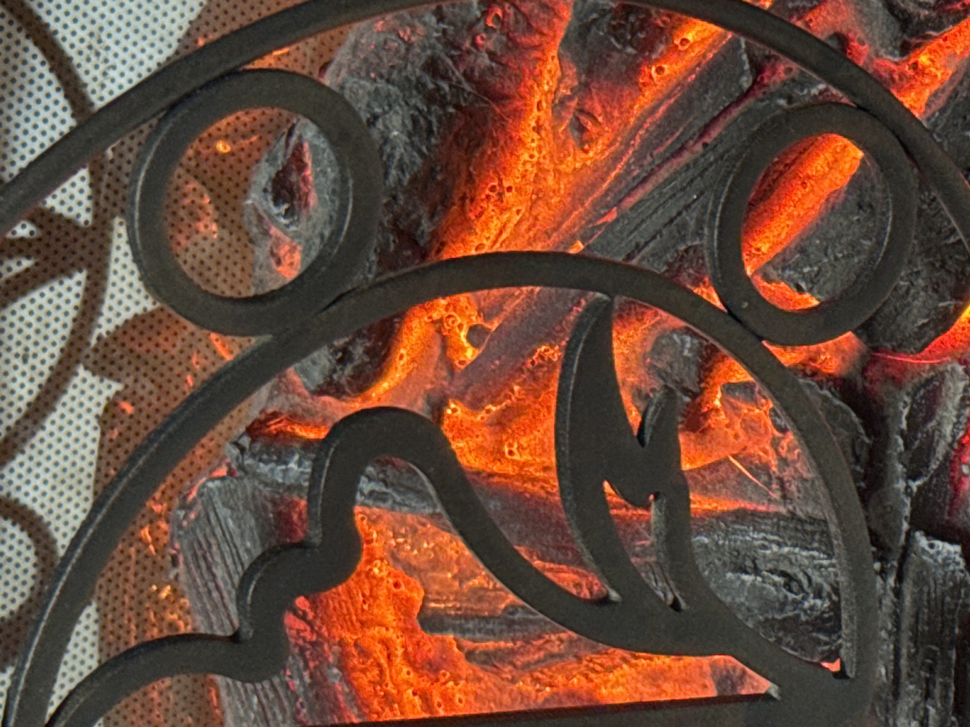 The image displays a close-up view of a decorative electric fireplace insert. The insert features faux logs that glow with a vibrant orange and red light, simulating a real fire. A dark metal decorative screen with swirling vine-like patterns partially obscures the glowing logs, adding an aesthetic element to the design. The background of the insert appears to be a textured surface with a subtle dotted pattern. The overall impression is one of warmth and a simulated fireplace ambiance. There are no people or specific geographical features that would indicate a location in Sinyavino, Russia.