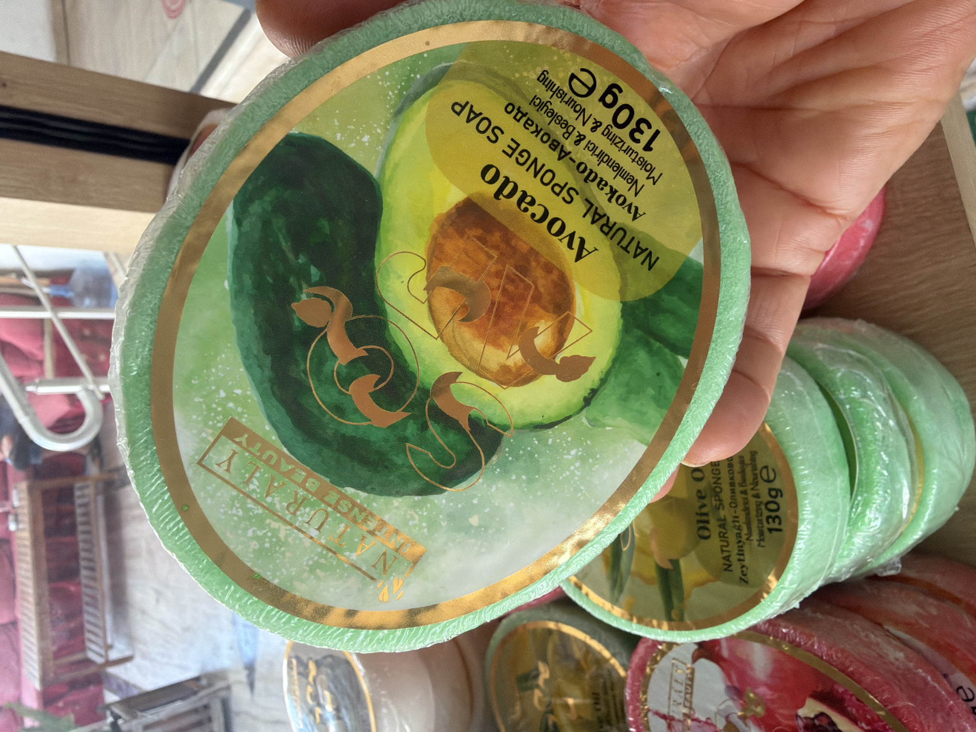 In Marmaris, Turkey, a close-up shot captures a hand holding a circular, packaged natural sponge soap with an avocado design. The soap is green and has a golden border with the brand "NATURALLY INTENSE BEAUTY" printed in gold letters. The front of the package displays an illustration of an avocado cut in half, with text indicating "Avocado NATURAL SPONGE SOAP" and "Moisturizing & Nourishing." Below this, it specifies "130g e." In the background, other similarly packaged soaps are visible, including one with a pomegranate design. The retail environment features wooden shelving and furniture, suggesting a shop setting.