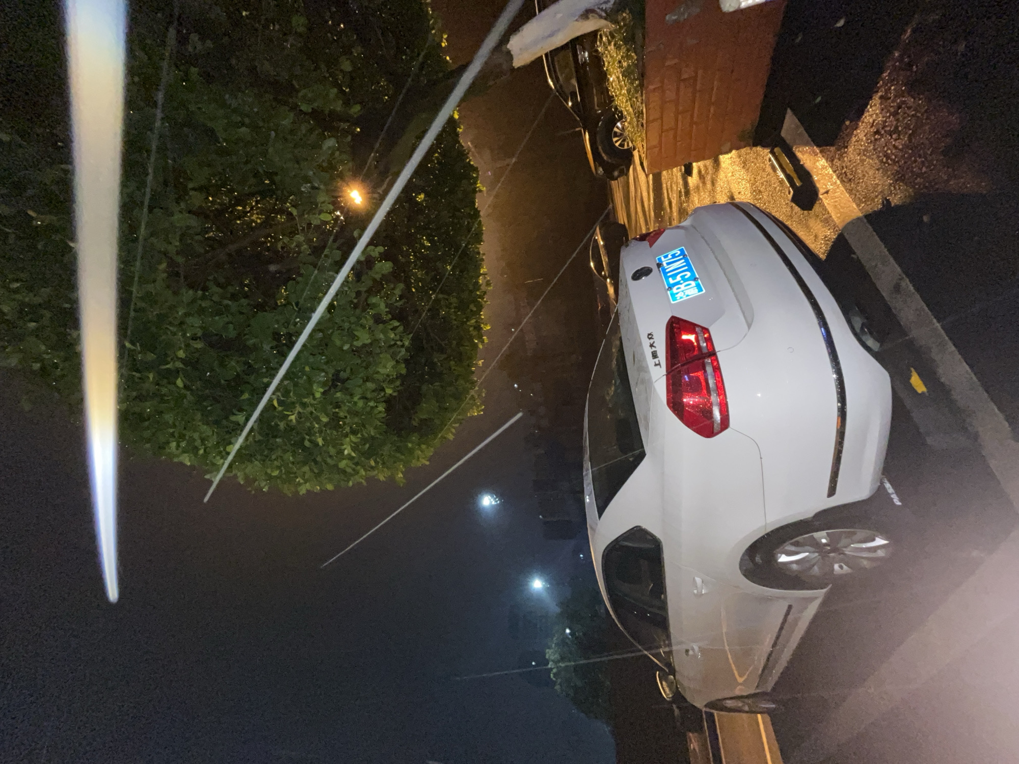 The image depicts an outdoor night scene within the urban environment of Jiulongpo District, Chongqing, China. A white Volkswagen Passat sedan is prominently positioned in the foreground, parked on a wet, asphalt surface. The car's rear, angled slightly towards the left, displays a blue Chinese license plate bearing the registration "渝B·51N75" and "上海大众" (Shanghai Volkswagen) badging on the trunk lid. A yellow-and-black wheel stop is visible behind the vehicle's rear bumper.
The asphalt ground is notably wet and reflective, indicating recent precipitation or damp conditions, with segments of white parking bay markings visible. A large tree with dense green foliage occupies the right side of the frame, partially obscuring the background. Through the foliage, a single yellow-orange streetlight emits a distinct glow. The dark night sky above is traversed by multiple elongated, bright streaks of light, which appear to be lens flares or reflections from intense artificial light sources.
In the mid-ground, to the right of the white sedan, two dark-colored vehicles are parked, one identifiable as a black SUV. Further in the background on the left, several distant, unidentifiable white and bluish lights are visible, suggesting other light sources or structures. A reddish-orange brick planter box is partially visible in the far right foreground. No individuals are present in the scene. The license plate prefix "渝B" confirms the vehicle's registration within Chongqing Municipality, aligning with the specified location of Jiulongpo District.