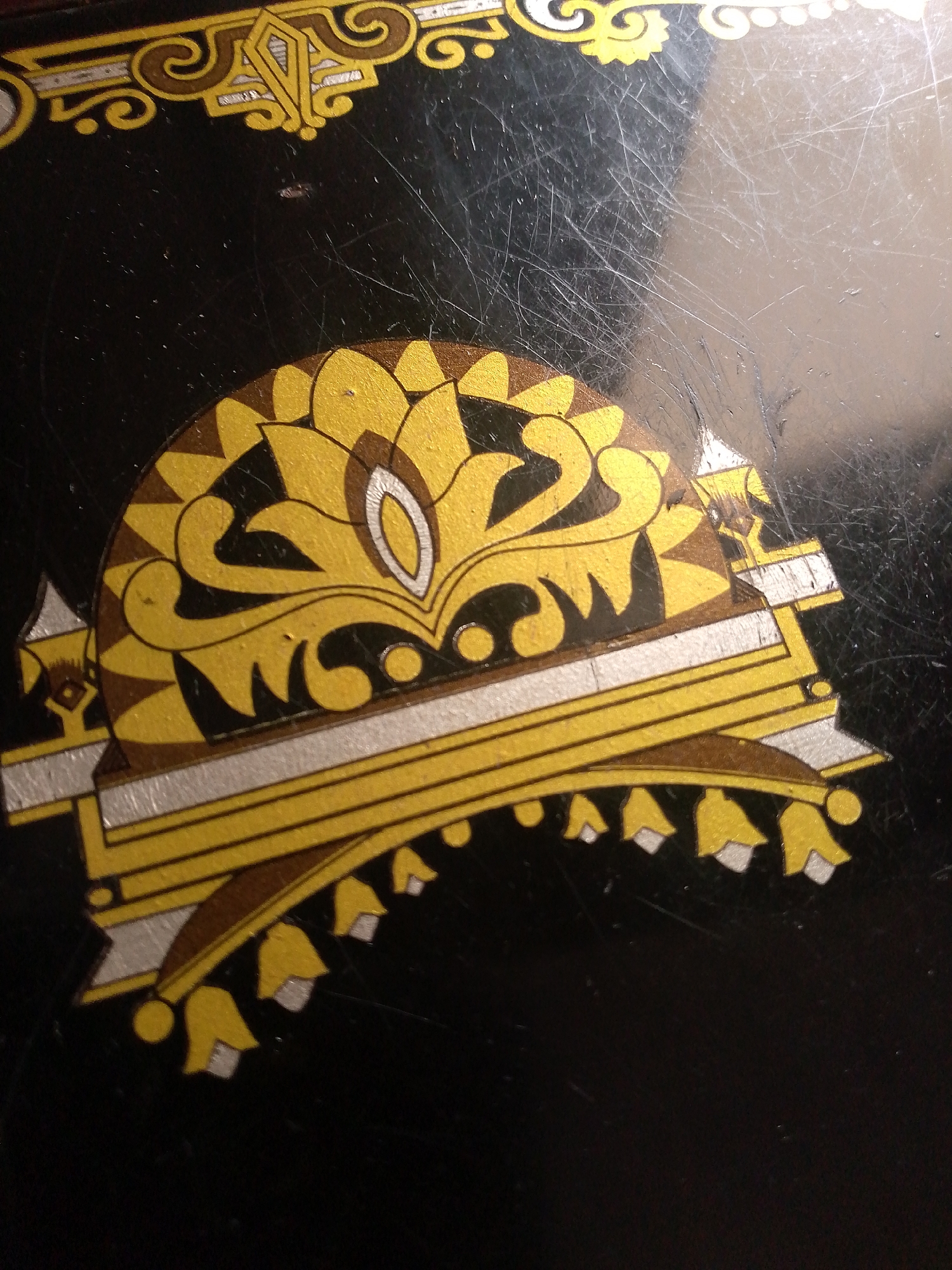 The image displays a close-up view of a decorative element on a black surface, likely an antique sewing machine or similar item. The decoration features intricate patterns in gold, brown, and silver. A prominent semicircular motif at the bottom is composed of stylized floral or fan-like shapes in gold, outlined with black and accented with white details. This motif is bordered by parallel lines of silver and gold, with a scalloped edge of smaller gold shapes accented with silver. Above this, a more complex design incorporates swirling golden elements and geometric shapes. The black surface exhibits numerous fine scratches, indicating age and wear. The scene is devoid of any people or immediate context that would suggest a location within the city of Jauro Yinu, Nigeria. The image focuses solely on the decorative artwork.