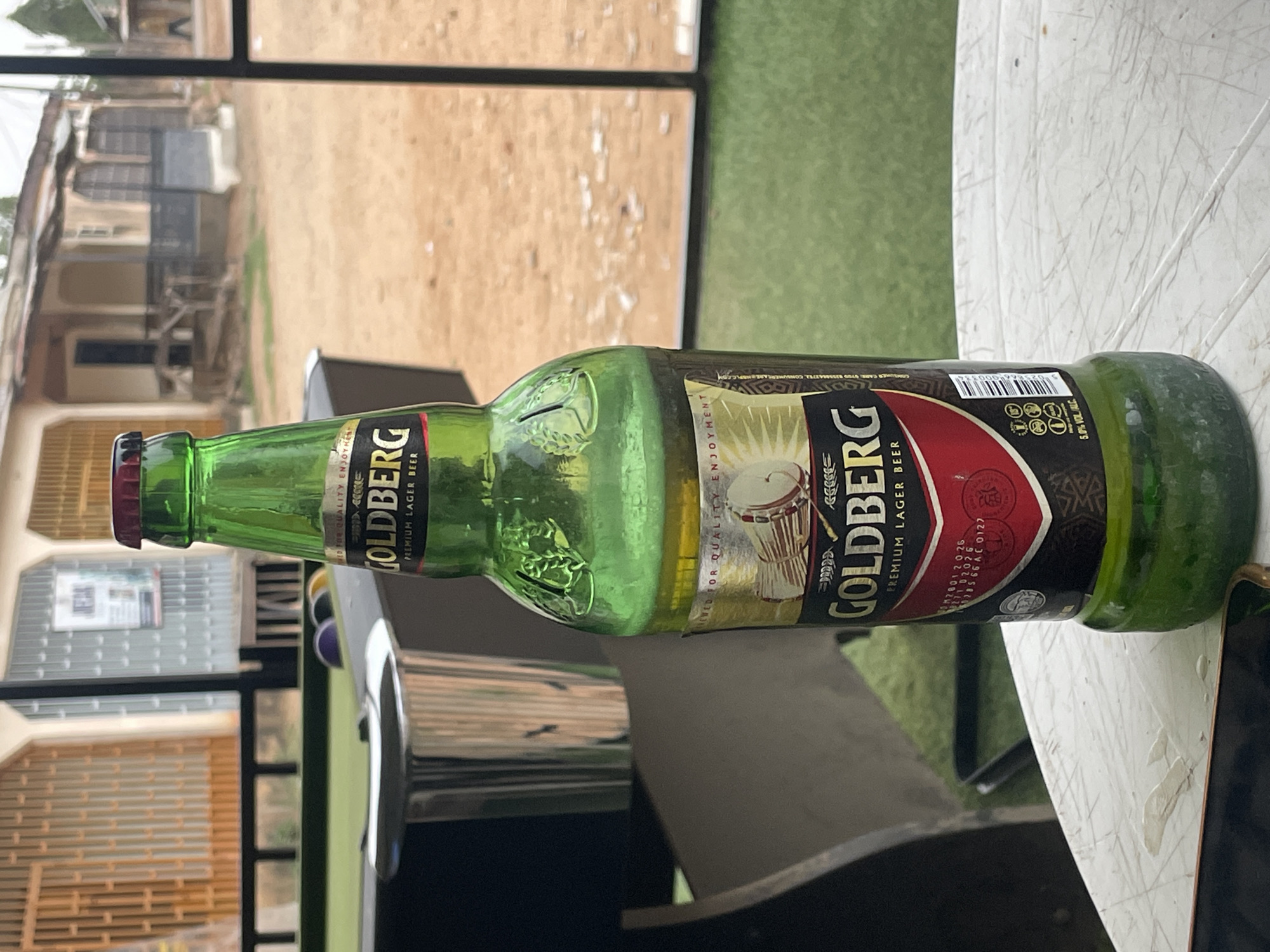 A green Goldberg Premium Lager Beer bottle lies horizontally on a white, weathered table. The bottle label features the "Goldberg" logo, an image of a drum, and the phrase "Premium Lager Beer." The bottle cap is red.
The background includes a blurred view of a building with light-colored walls and a brown roof, suggesting a residential area. A metal frame structure partially obscures the view. A patch of green grass is also visible.
The scene appears to be outdoors, possibly on a patio or veranda. It's not possible to confirm the location in Jos, Nigeria, without further context or visual clues specific to that city.