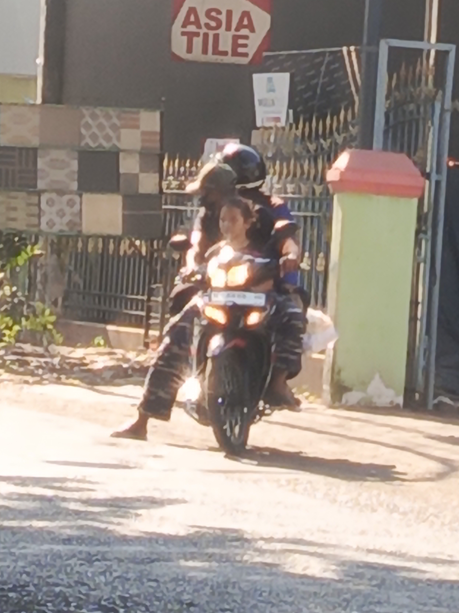 In Sidowungu, Indonesia, a person wearing a dark helmet and a patterned shirt rides a motorcycle. They are seated behind another individual who is wearing a dark helmet and a blue shirt. The passenger is holding onto the driver. The motorcycle has a visible license plate and illuminated headlights. They are on a paved road, with a green wall and a metal gate visible in the background. A sign with "ASIA TILE" written on it is also present. The environment suggests a commercial or residential area.