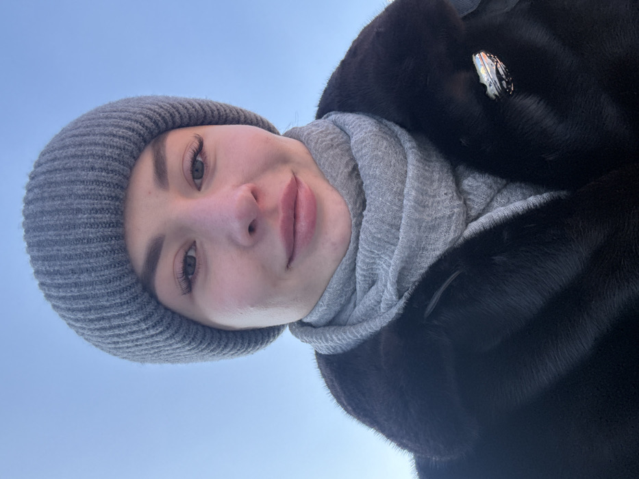 The image displays a head-and-shoulders portrait of a single female individual centered against a clear blue sky. She possesses light skin and is looking directly into the camera with a subtle, closed-mouth smile. Her attire consists of a grey ribbed knit beanie, a grey textured scarf wrapped around her neck, and a dark, short-pile fur coat. A metallic, multi-faceted brooch is pinned to the right lapel of the fur coat. The background is an unobstructed, uniformly light blue sky, indicating an outdoor environment during daylight. No specific landmarks or architectural features are visible to determine a precise location within Kyiv, Ukraine. The heavy winter clothing suggests cold weather conditions.