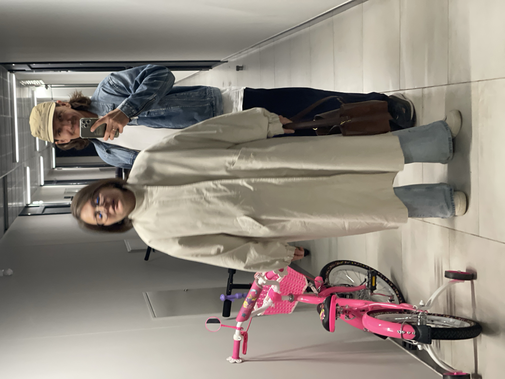 Two individuals are visible in an interior space with a pink bicycle nearby. The first individual, possibly male, is wearing a light-colored baseball cap and a blue denim jacket. They are holding a smartphone and appear to be taking a picture. The second individual, likely female, is wearing glasses and a long, light-colored coat. Both individuals are standing in a hallway or corridor with tiled flooring and white walls. The pink bicycle has a white training wheel assembly attached to it. The location is undetermined, as no specific city or location identifiers are apparent.