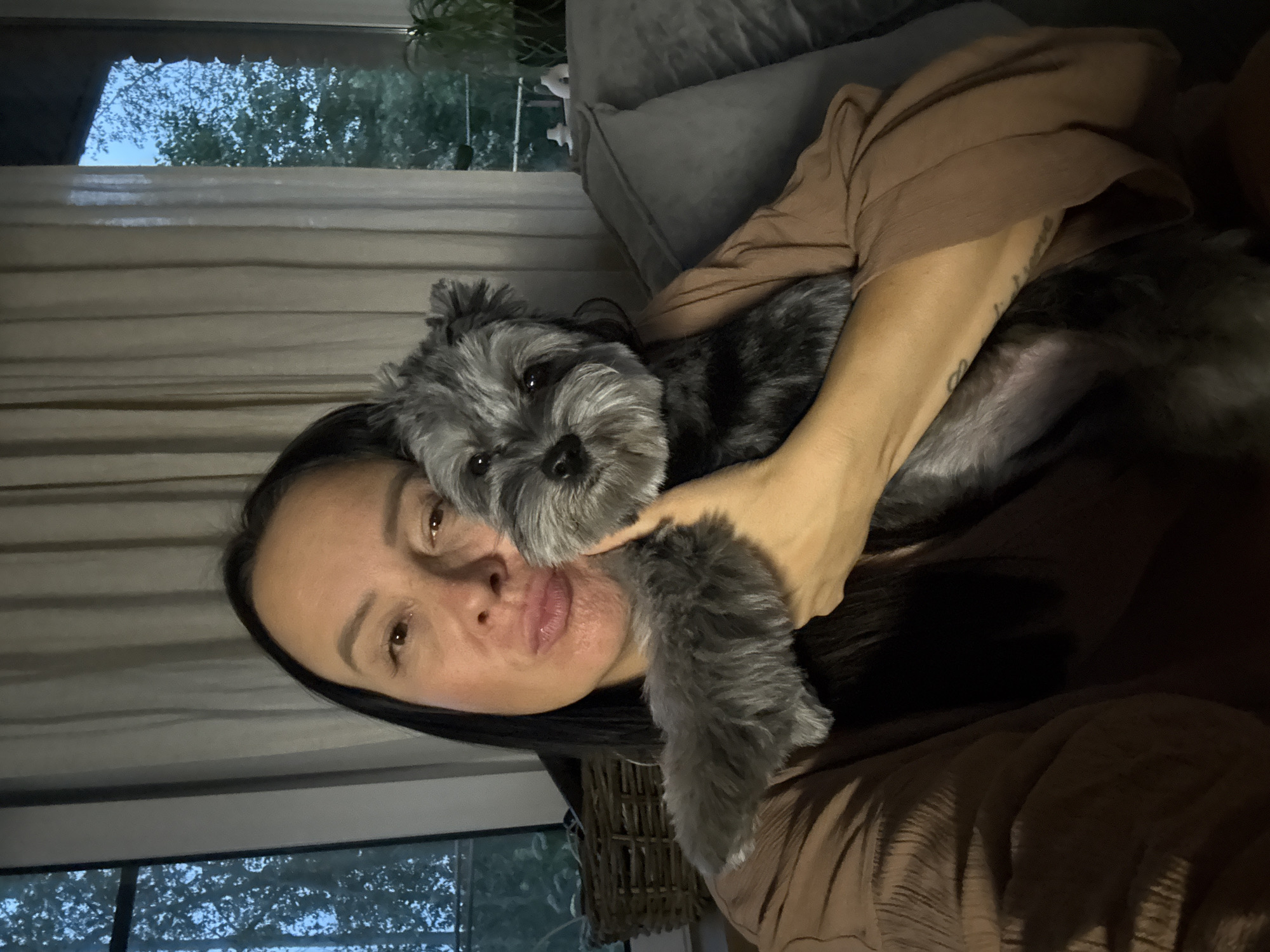A woman is taking a selfie while holding a small, grey dog. She has dark hair and is wearing a brown top. The dog has grey fur and is looking at the camera with its head tilted. They are indoors, likely in a living room, as a couch and a window with trees visible outside can be seen in the background. The lighting suggests it is either early morning or late evening. The woman's expression is relaxed, and the dog appears content. There is a tattoo visible on the woman's arm.