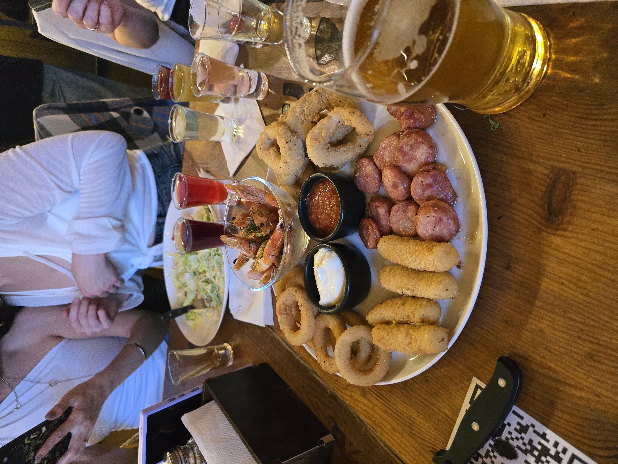 The image depicts an indoor dining and socializing scene at an establishment in Vologda, Russia. The primary focus is a wooden table laden with food and drinks.
In the foreground, a large white oval platter is central, containing a variety of fried appetizers: approximately seven golden-brown onion rings, seven breaded mozzarella sticks, and numerous slices of grilled, reddish-brown sausages. Two small black bowls on the platter hold dips: one white and creamy, and one reddish and chunky. To the left of the platter, a glass bowl contains cooked shrimp garnished with green herbs.
To the right, a clear glass mug holds light-colored beer with a foamy head. In the mid-background, six shot glasses contain various colored liquids, including red, clear, yellowish-orange, and pinkish. Another small shot glass with amber liquid is visible on the far left. A white plate with a green salad is partially visible in the left background, next to another small dish.
Four individuals are partially visible around the table. On the far left, a woman in a white, one-shoulder top is looking at a dark smartphone held in her hands. A silver necklace with a heart pendant is visible. Behind her, another person in a white, tied-front top is seen, with their hands gesturing or adjusting clothing. A partial view of a person in a dark t-shirt and jeans is in the center-right background, and an arm with a ring on a finger is visible on the far right.
On the bottom-left, a black folding knife rests on a piece of white paper displaying a QR code. A black container holding white paper napkins and a small silver shaker are also on the left side of the table. The table itself is dark wood with visible grain. The lighting suggests an evening or indoor ambiance.