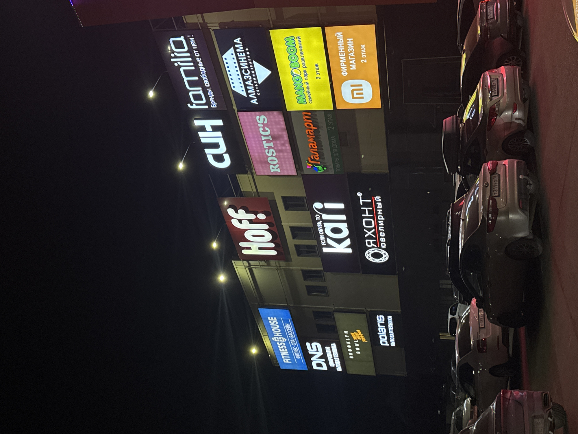 The image shows a nighttime scene with a multi-story commercial building featuring numerous illuminated signs, likely a shopping or entertainment center. The signs, stacked vertically, display various brands and stores including "Familia," "CUH," "Hoff," "Fitness House," "DNS," "Polaris," "Rostic's," "Almaz Cinema," "Mango Boom," "Kari," and "Yakht." Several parked cars are visible along the right side of the frame. Based on the branding and signage language (Russian), it is likely located in Russia. Given the logos and information, it is possibly a shopping center in Izhevsk, Russia.