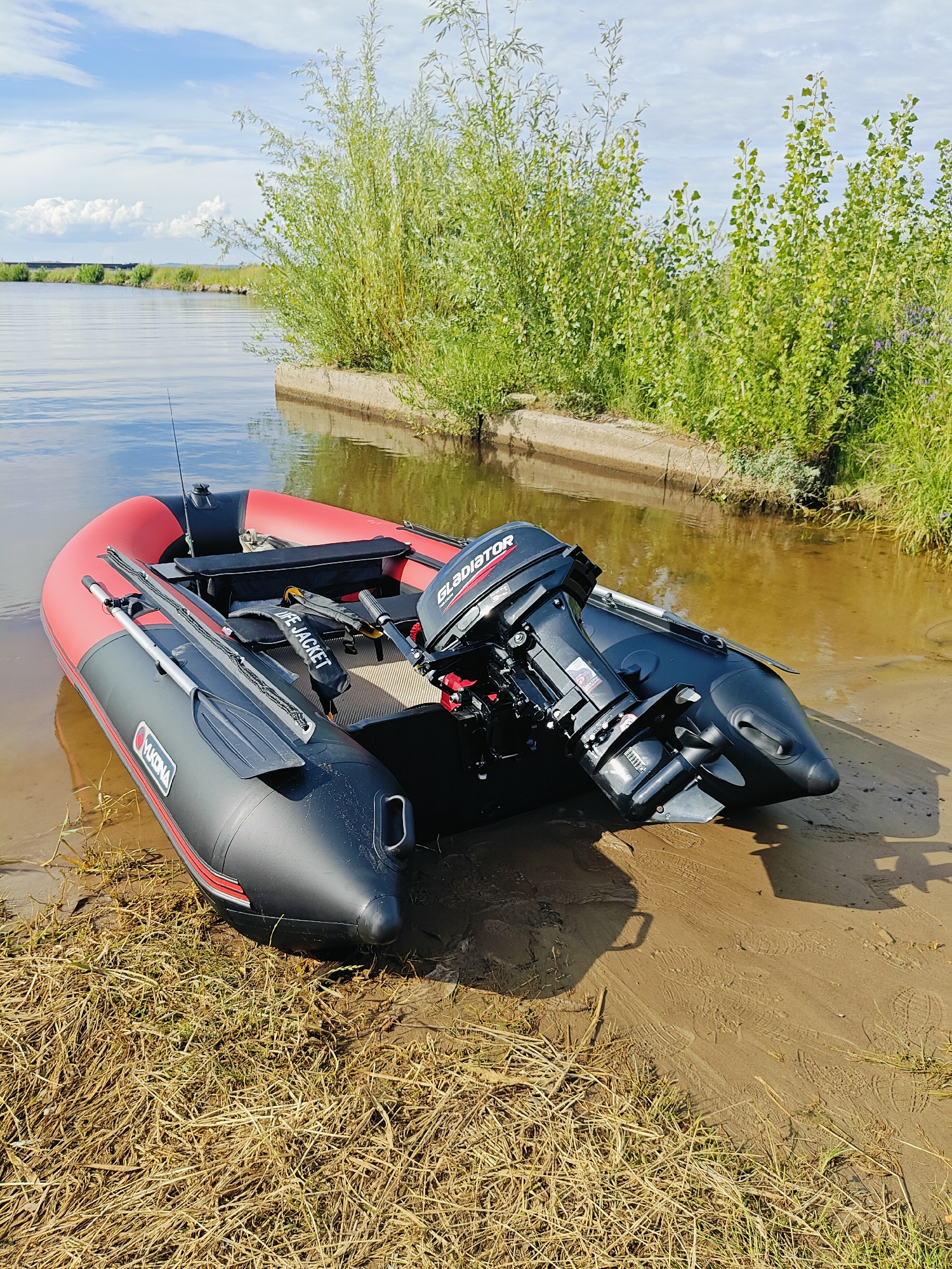This media file depicts a red and black inflatable boat with an outboard motor resting on a sandy shore next to calm, reflective water. The boat is positioned at an angle, with its bow pointing towards the left and its stern, containing the motor, to the right. The motor has "GLADIATOR" visible on its casing. Oars are attached to the sides of the boat. A life jacket is also visible inside the boat. The shore is a mixture of sand and dry grass.
Behind the shore, lush green vegetation, including tall, bushy trees and smaller plants, grows along a concrete embankment. The water reflects the blue sky, which is dotted with fluffy white clouds. The overall scene suggests a calm, sunny day, likely in the late morning or early afternoon given the bright lighting and clear sky. There are no people visible in the image. The presence of the boat and the water implies a recreational setting, possibly for fishing or water sports.
Visible text includes "GLADIATOR" on the motor and "YUKONA" on the side of the inflatable boat. The location context is Chaikovsky, Russia.
