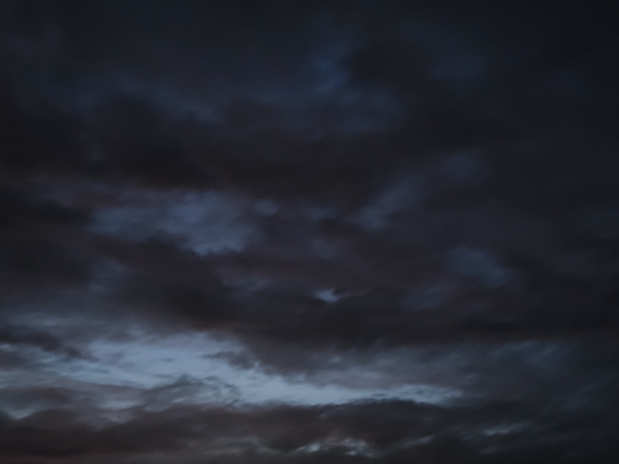 An ominous, dark sky filled with heavy, bruised clouds dominates the frame, with hints of pale blue light peeking through the gloom. The clouds are textured and layered, appearing dense and potentially threatening, suggesting an impending storm or the aftermath of one. There are no discernible objects, people, or environmental features within the frame, making the sky the sole subject of the image. The location is identified as Unknown, Unknown.