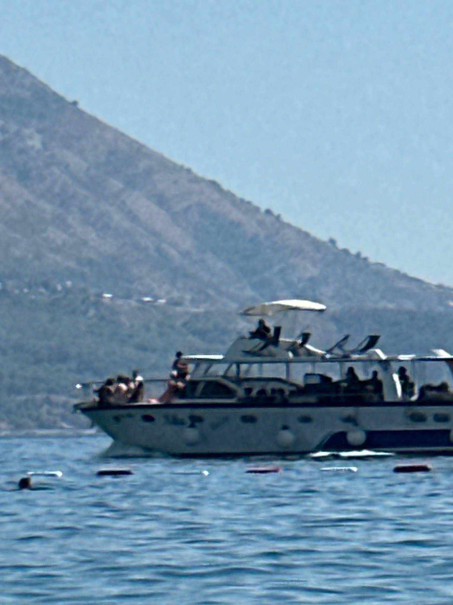 A wide shot captures a boat on the water with a mountainous backdrop, likely near Boreti, Montenegro. The boat, a leisure vessel, appears to be carrying several passengers, visible as silhouettes against the sunlight. Some individuals are standing on the deck, while others are seated. The water is a deep blue, with gentle ripples indicating calm conditions. In the distance, a rugged mountain slopes down towards the water, its surface textured with vegetation and possibly some scattered structures. The sky above is a clear, pale blue. Several buoys are floating on the water's surface, some red and white, others plain white, suggesting a marked area or a navigational aid.