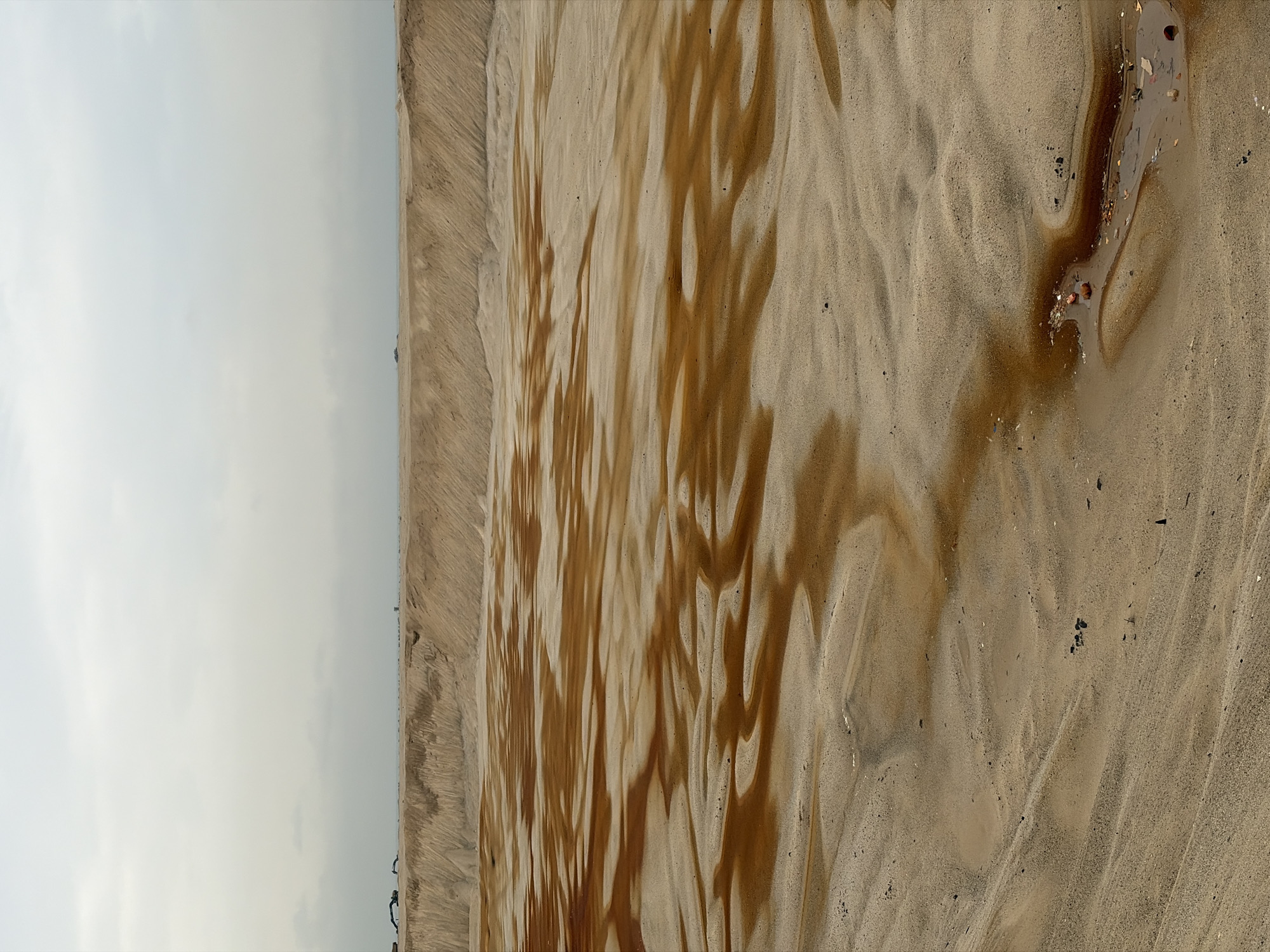 The image depicts a sandy landscape with some damp areas stained reddish-brown, possibly from iron-rich water. The terrain is uneven, with undulating mounds and shallow depressions. In the foreground, there is a small pool of water with some debris, including what appears to be fragments of pottery or brick. The background features a large, sandy embankment, and in the distance, a hazy horizon suggests a body of water or a flat, distant landmass. The sky is overcast and pale. The scene suggests an open, potentially industrial or construction site, given the presence of what appear to be construction vehicles or equipment on the distant embankment. No people are visible in the immediate scene. The location is identified as Alaguntan, Nigeria.