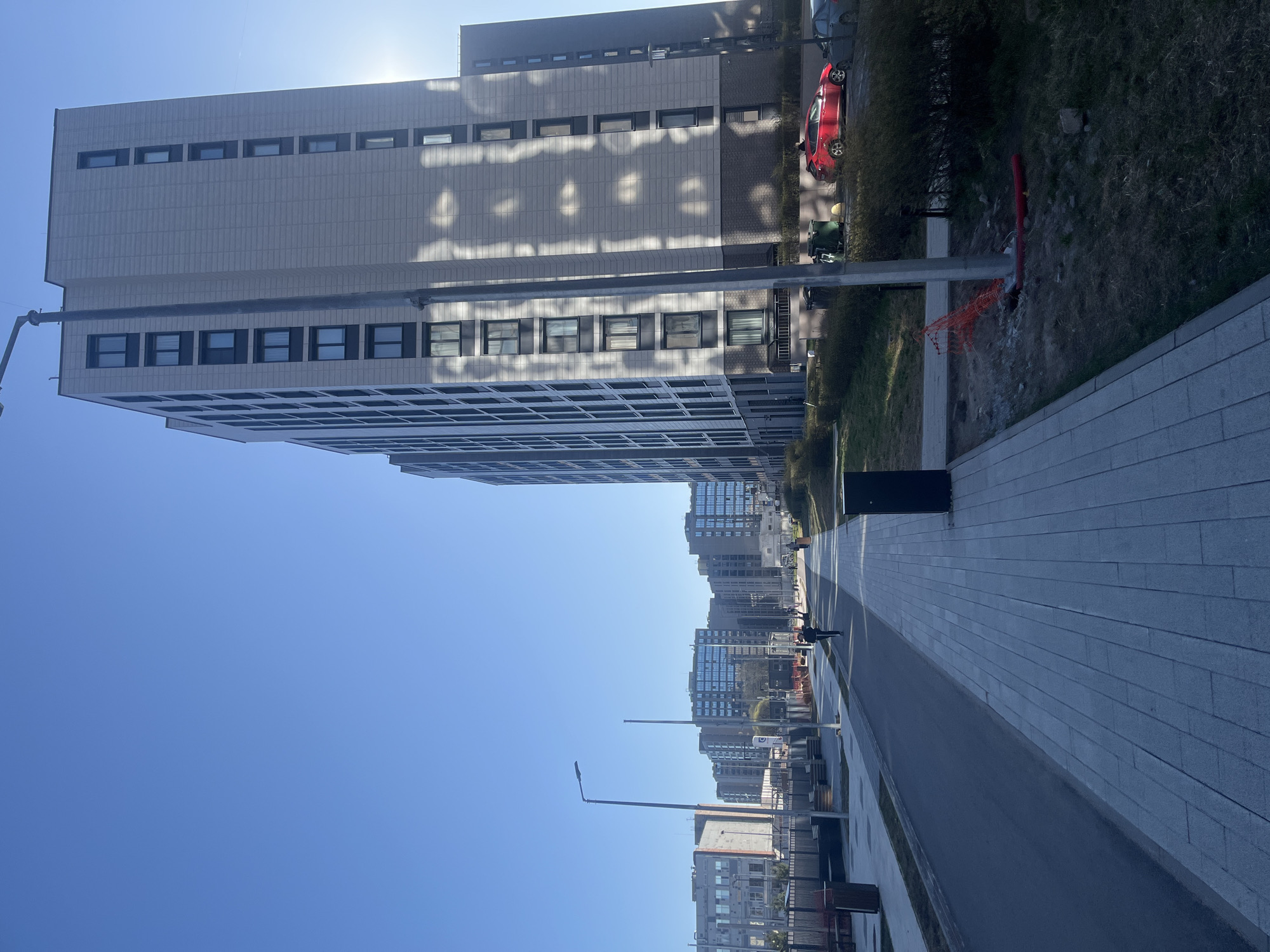 The image depicts a bright, sunny urban street scene in Saint Petersburg, Russia. A tall, modern building with a light-colored facade and numerous dark-framed windows dominates the left side of the frame. Sunlight reflects off its surface, creating abstract patterns, and the sun itself is visible as a bright flare behind the building's top edge.
A wide, paved area, comprising a light gray stone walkway and a darker asphalt road, stretches into the distance on the right. A prominent streetlight stands in the foreground. Further down the street, more contemporary buildings are visible, and a lone figure can be seen walking on the pavement.
On the ground level, alongside the main building, a red car is parked amidst green grass and low bushes. Nearby, orange safety netting and a red pipe protruding from the ground suggest ongoing construction or maintenance work. The clear, bright blue sky indicates fair weather, and the strong sunlight casts shadows, suggesting it is either morning or afternoon. No discernible text is visible in the scene. The overall impression is one of a quiet, modern city environment on a clear day.
