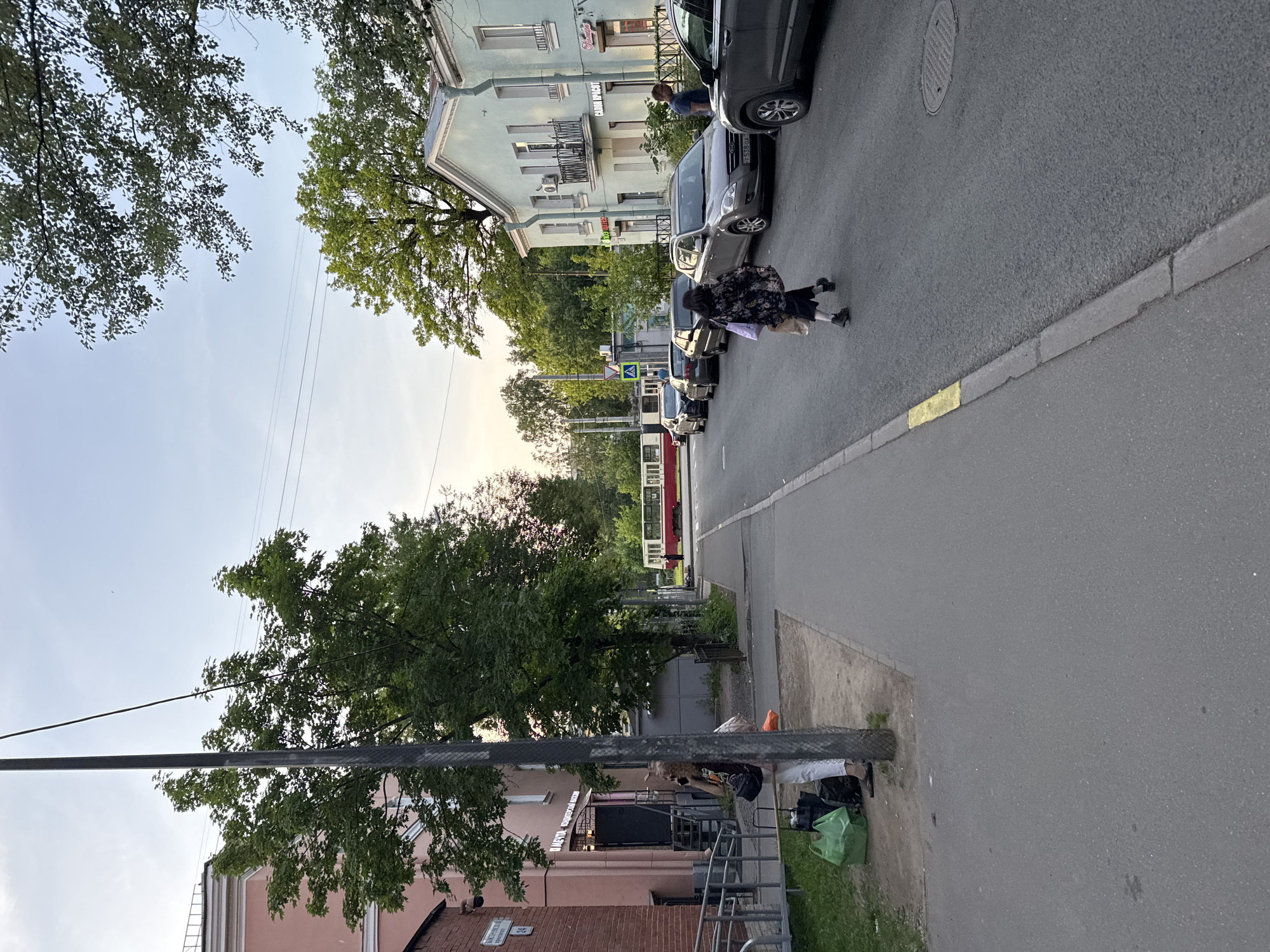 The image depicts a street scene in Saint Petersburg, Russia, likely during the late afternoon or early evening, given the lighting. On the left, a pink building with a brick facade is visible, with the number "24" and a sign that appears to read "МЕЧТА - КОНДИТЕРСКИЙ МАГАЗИН" (Dream - Confectionery Store). A person with curly hair is standing on the sidewalk, talking on a mobile phone, with luggage including green shopping bags and black bags at their feet. Further down the sidewalk, a light blue building with balconies and signs that say "САЛОН КРАСОТЫ" (Beauty Salon) is present.
The street is lined with parked cars, including a dark grey sedan in the foreground on the right. Several other cars are parked sequentially on the right side of the road. Across the street, a tram is visible with a red and white color scheme. People are walking on the sidewalk and crossing the street. The environment is urban, with trees lining the street and a pale sky overhead. The road surface is asphalt, with a yellow painted line at the edge of the sidewalk in some areas.