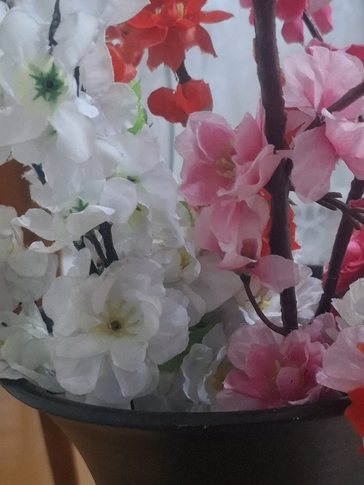 The image shows a close-up of artificial flowers in a dark vase. The flowers are primarily white and pink, with some red accents visible. The white flowers are clustered together and have delicate, layered petals with a yellow center. The pink flowers are also multi-petaled and appear to be on a branch, with darker brown stems. Some green foliage is interspersed among the blooms. The vase is dark, possibly black or a deep brown, and has a wide opening. The background is a soft, out-of-focus blueish-gray, suggesting a wall or a window. There are no people or animals present. The lighting is soft, with no harsh shadows, indicating an indoor setting. The image does not provide any visual cues to pinpoint its location within the city of Almaty, Kazakhstan, or any specific urban environment.