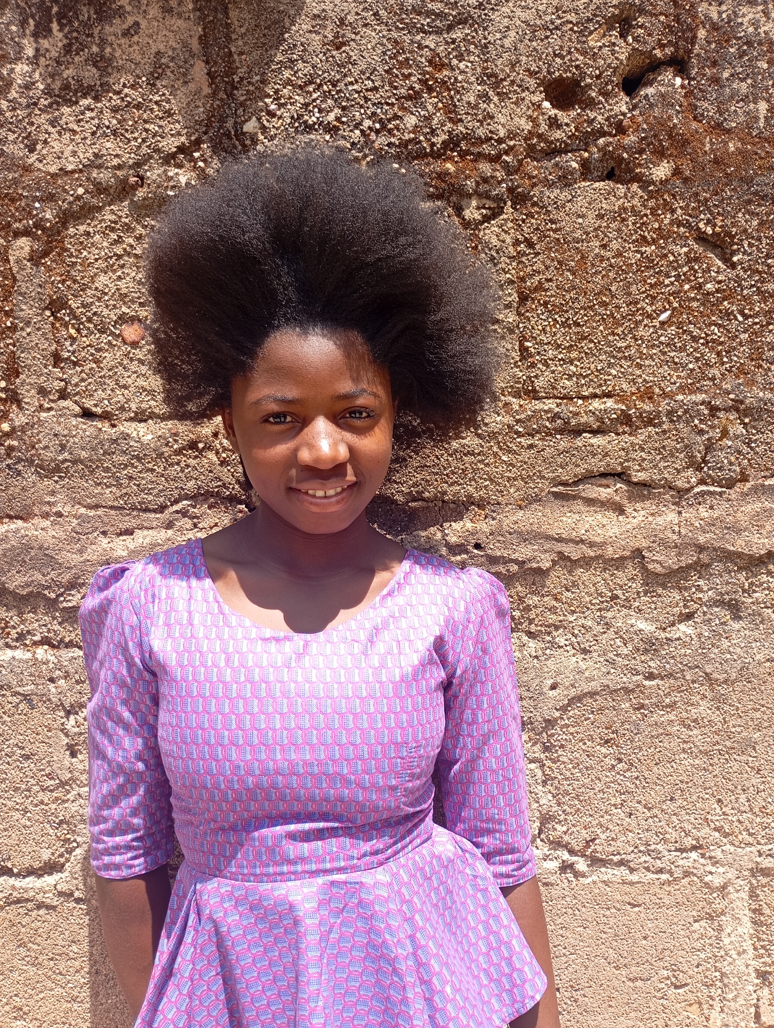 A young, dark-skinned woman with a large afro hairstyle is standing outdoors. She is wearing a purple and white patterned dress. She is smiling and looking directly at the camera. Behind her is a textured, tan-colored wall. The lighting suggests a sunny day. This location is the city of Jos, Nigeria.