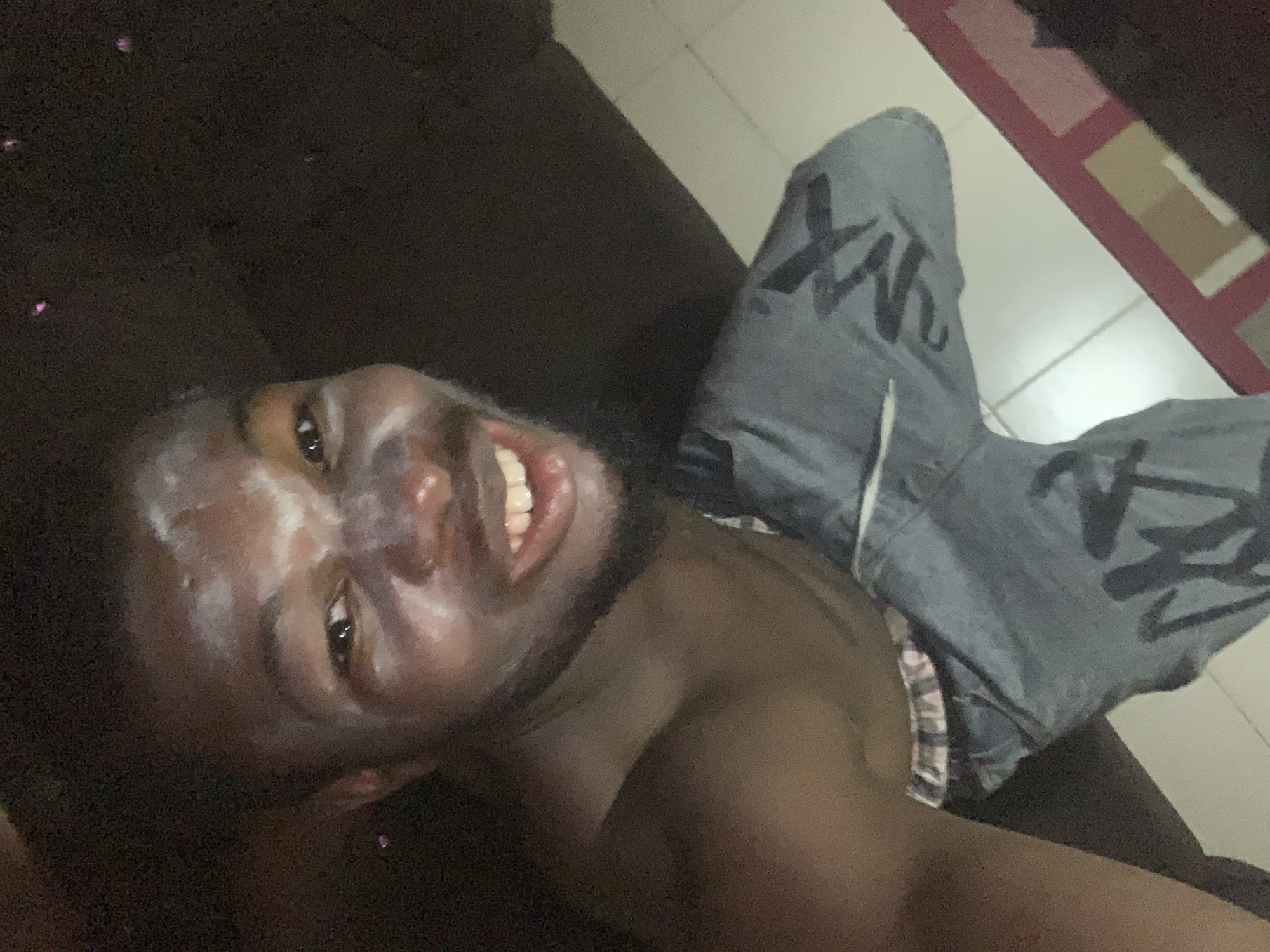 The media file features a young Black man lying down, likely on a dark brown couch or bed, with a broad, joyful smile revealing his teeth. He has white or light-colored substance, possibly paint or powder, smeared across his forehead, nose, and cheeks, suggesting it might be part of a celebration, ritual, or artistic expression. He appears shirtless, exposing his shoulder and upper chest.
He is wearing light blue jeans adorned with large, dark, graffiti-style letters, prominently displaying "XK" on his left leg and similar, partially obscured letters on his right leg. A piece of checkered or striped fabric, possibly his underwear or the inner lining of his clothing, is visible near his hip.
The setting appears to be indoors, likely a residence in Onitsha, Nigeria, given the provided context. The floor is covered with light-colored tiles, possibly white or cream, featuring a pattern of red and tan/brown squares or lines along the edge, which might be a rug or floor design. The lighting is dim, indicating it could be evening or a poorly lit room. The man's happy expression suggests a positive and possibly festive mood.