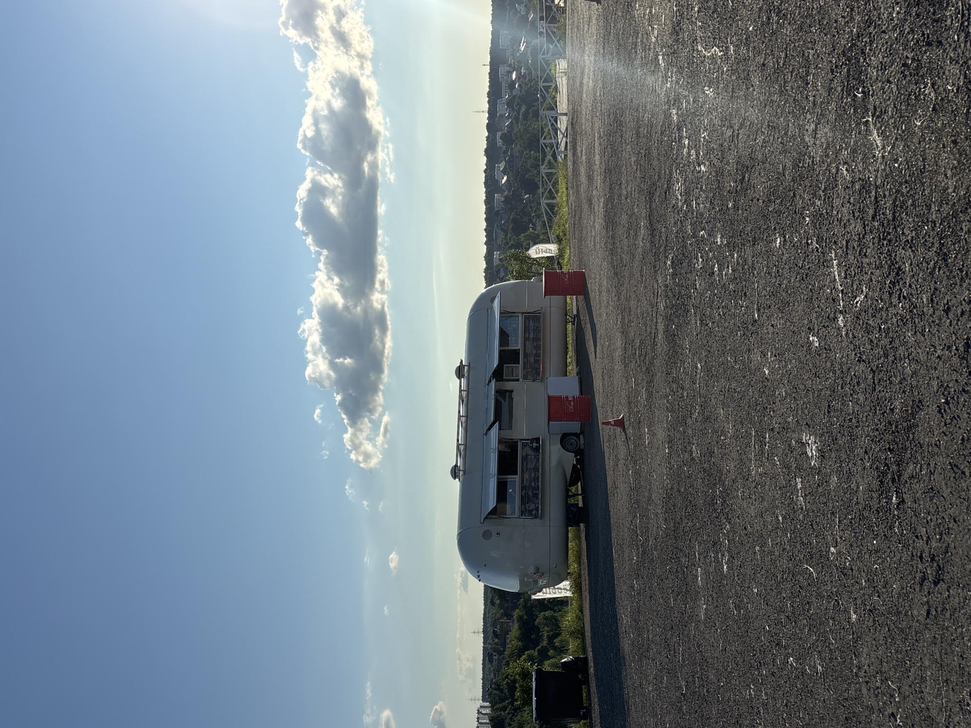 An Airstream-style food truck is parked on a gravel lot with a scenic overlook in the background. The food truck is white with open windows showcasing menus. A red trash can and a red barrel are near the truck, as well as a traffic cone. The background features a lush green landscape with buildings and trees, under a bright blue sky with scattered clouds. The location is identified as Romashkovo, Russia.