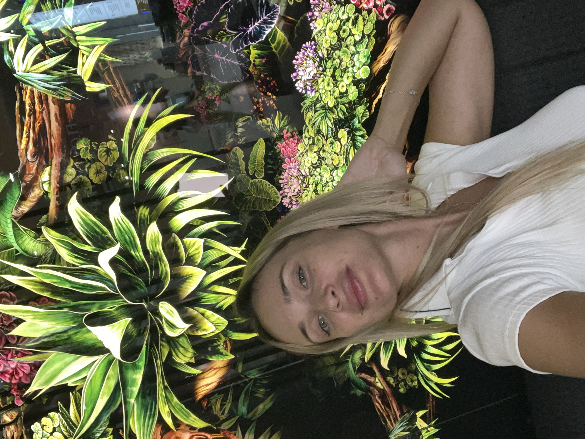 A young woman with blonde hair and green eyes takes a selfie in front of a vibrant floral mural. She is wearing a white collared shirt. Her left arm is bent, and her hand is resting on the back of her head. The mural features large green leaves, pink and purple flowers, and brown branches. The background appears to be a dark, possibly reflective surface, with some indistinct elements suggesting an indoor setting. The image appears to have been taken in Odesa, Ukraine.