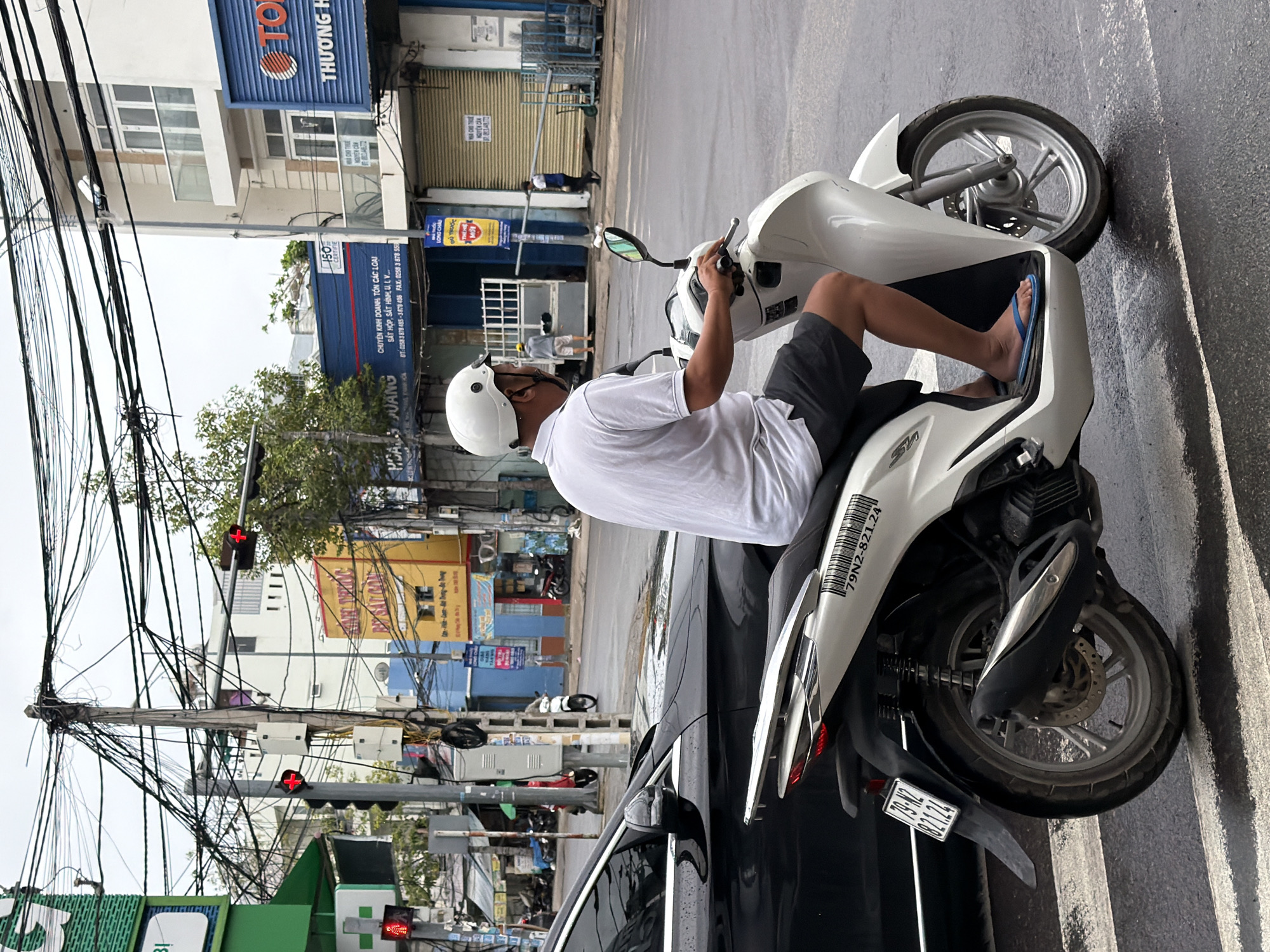 The image depicts a street scene in Nha Trang, Vietnam, likely during daylight hours with overcast skies. A man wearing a white helmet, white short-sleeved polo shirt, and dark shorts rides a white scooter, identified by the license plate "79N2-821.24". He is positioned in the foreground, with his right hand on the scooter's handlebars and his left hand visible. The scooter is on a wet asphalt road marked with white pedestrian crossing lines, indicating an intersection.
To the left of the scooter, a portion of a black car is visible. The background reveals a city street with various buildings. One building on the right has a blue facade with the word "THƯƠNG H" visible, suggesting a business. Another building further back is yellow with Vietnamese text. A significant feature of the urban environment is a prominent utility pole heavily laden with a dense network of black electrical wires and cables, creating a cluttered overhead appearance. Traffic lights are visible on the utility pole, with one red light illuminated. Other elements in the background include other vehicles, storefronts with rolling metal shutters, and what appear to be scaffolding or construction materials being transported by individuals. The overall impression is a busy urban street with a mix of traffic, commerce, and utilitarian infrastructure.