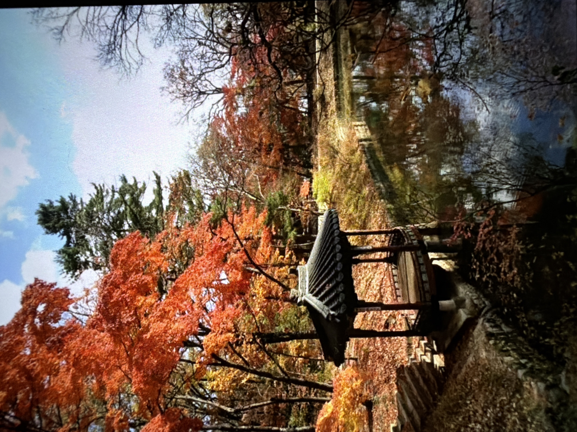 The image depicts a tranquil scene in Ungasan, Indonesia, during autumn. A traditional wooden pavilion with a tiled roof stands amidst vibrant foliage, with trees displaying hues of red, orange, and yellow. A narrow waterway or canal runs alongside the structure, reflecting the trees and sky. The atmosphere is peaceful, capturing the beauty of the fall season in this Indonesian location.