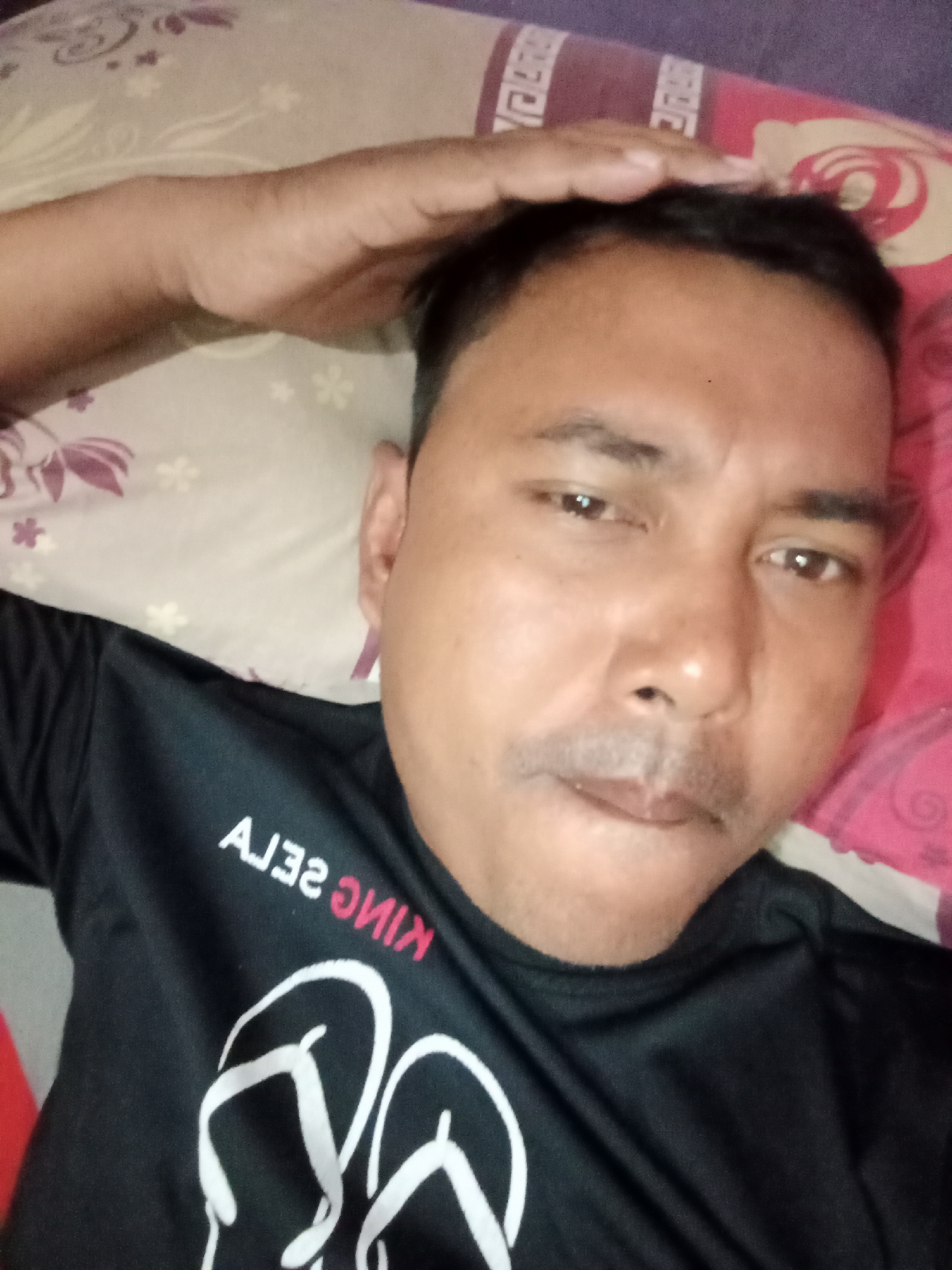 A selfie taken by a person lying on a bed. The person is wearing a black t-shirt with a graphic of flip-flops and text that appears to read "KING SEJA" in reverse. The background shows a patterned bedsheet with floral designs and a purple pillow. The image appears to be taken indoors. There is no identifiable information or context that would place this image in Bloro, Indonesia.