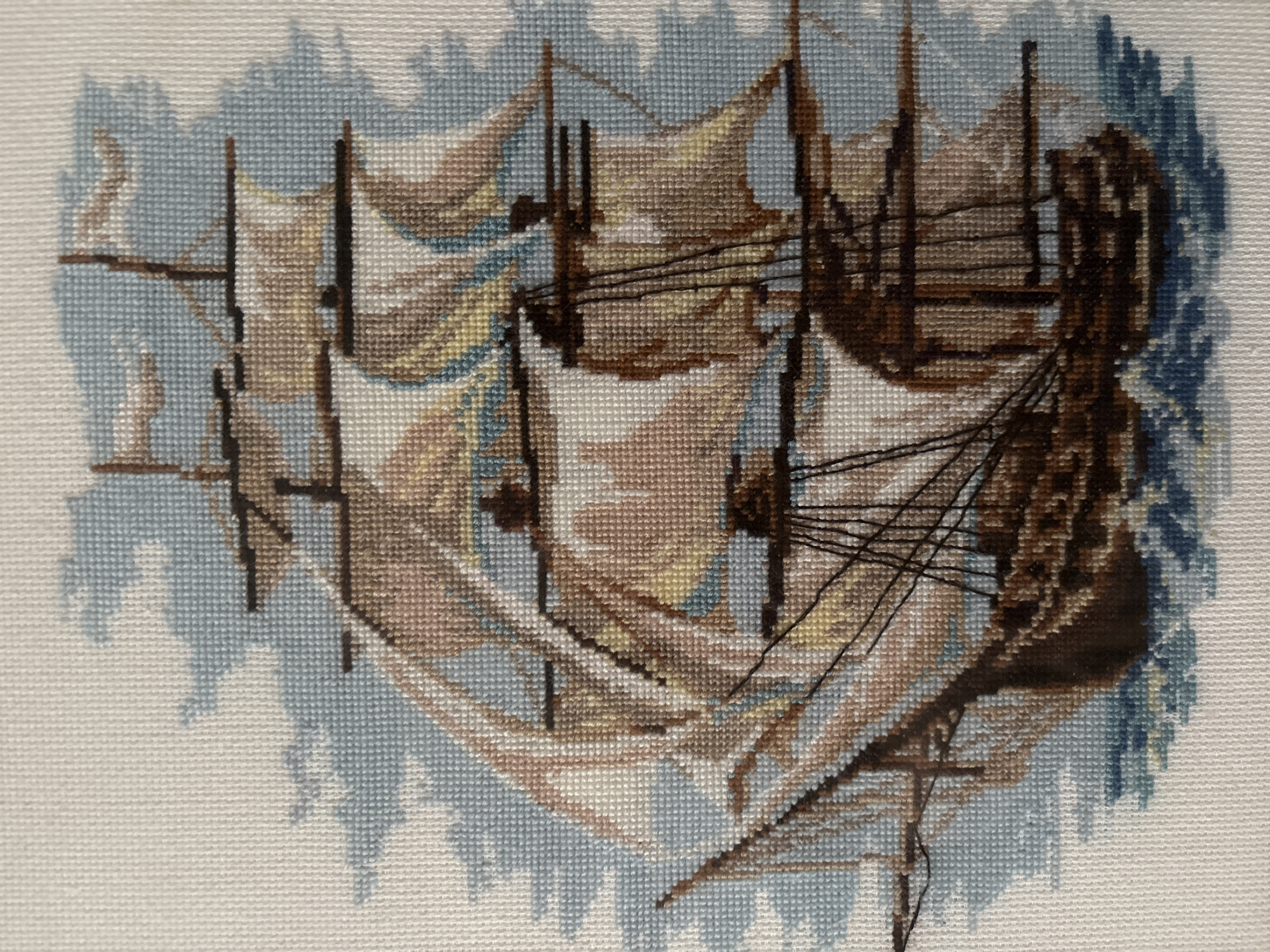 This is a cross-stitch embroidery of a tall ship sailing on choppy blue water. The ship has multiple masts and sails, rendered in various shades of brown, tan, and white, suggesting a dynamic and possibly windy scene. The background depicts stylized clouds in pale blue. The embroidery is on a white Aida cloth fabric, which is characteristic of cross-stitch.
There is no information in the image or its crops that suggests a specific location in Tomsk, Russia, or any other city. The scene is entirely fictional and depicted in the style of needlework.