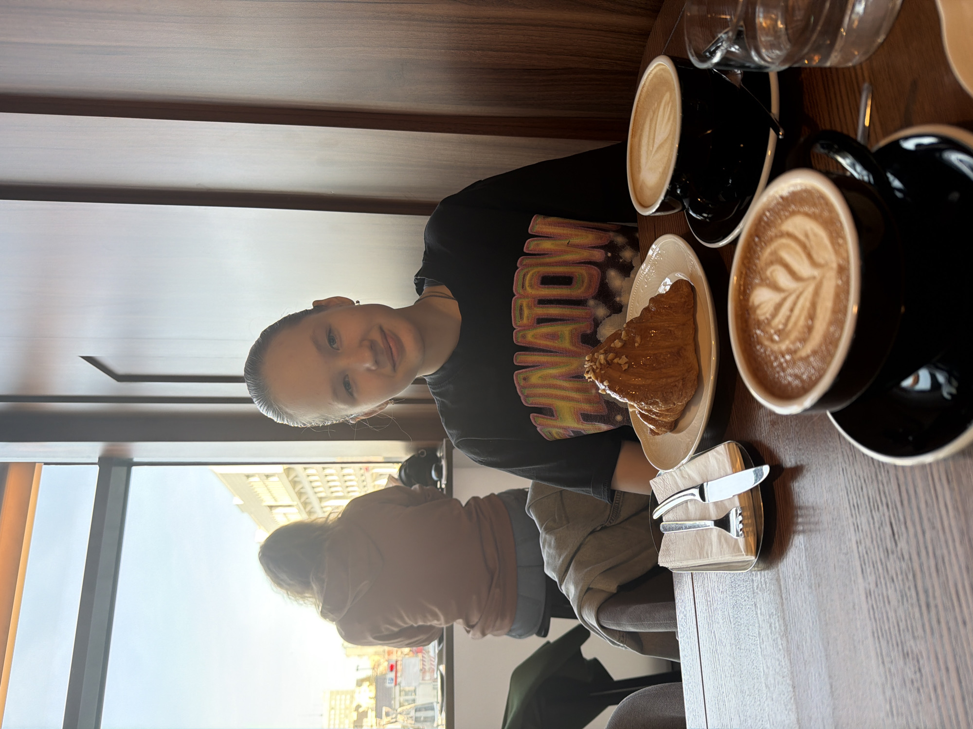 The image shows two people seated at a table, likely in a cafe or restaurant. One is a younger individual in a black t-shirt with a colorful "NATION" logo, positioned closer to the camera. The other is an adult facing away from the camera wearing a long sleeve shirt, lighter brown in color. The table is wooden. On the table, there is a croissant on a plate, cutlery, two black cups of coffee with latte art. There are also glasses of water in the background. A large window provides natural light, revealing a cityscape. Given the prompt, the location is Perm, Russia.