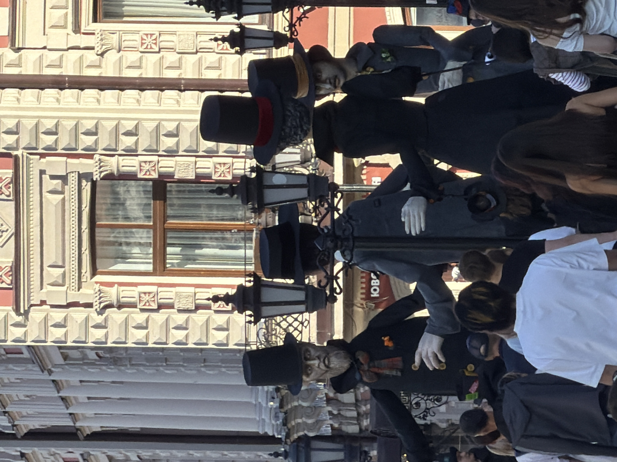 The image depicts an outdoor street scene in Kazan, Russia, specifically in front of the GUM Department Store, identifiable by its distinctive architectural style.
Four large, humanoid figures, appearing to be oversized puppets or mannequins, dominate the midground. All four are dressed in dark, formal, 19th-century style clothing, each wearing a top hat. From left to right:
1. A male figure with a grey beard, wearing a black top hat, a black coat with a yellow button, and a plaid red, orange, and blue scarf. His left arm is extended.
2. A male figure viewed from the back, wearing a black top hat with a red band and a black coat. This figure is integrated with a multi-headed black street lamp.
3. A female figure viewed from the back, wearing a black top hat with a golden-brown band and a long black dress or coat. This figure is also integrated with a multi-headed black street lamp.
4. A male figure, partially visible on the right, facing towards the right with his head slightly turned, wearing a top hat with a yellow-gold band, a grey coat with a green boutonniere, and faint facial hair.
The figures' arms are extended, some with white-gloved hands, as if in motion or interacting. The ornate black street lamps are a central feature, connecting the figures.
In the background, the multi-story GUM Department Store building features a light-colored facade with intricate details, including rusticated (diamond-cut) blocks, decorative columns with capitals, and patterned red and white floral or starburst motifs around windows. Several tall windows with brown frames and white curtains are visible. A red sign with white Cyrillic text, "ЮВОР" (YUVOR), is partially visible on a lower section of the building. Further left, columns of an adjacent or connected building are visible.
The foreground is occupied by a crowd of people, mostly seen from behind or partial profiles, wearing contemporary casual attire (t-shirts, hoodies). They appear to be observing the figures or navigating the street. The scene is brightly lit by daylight, casting some shadows.