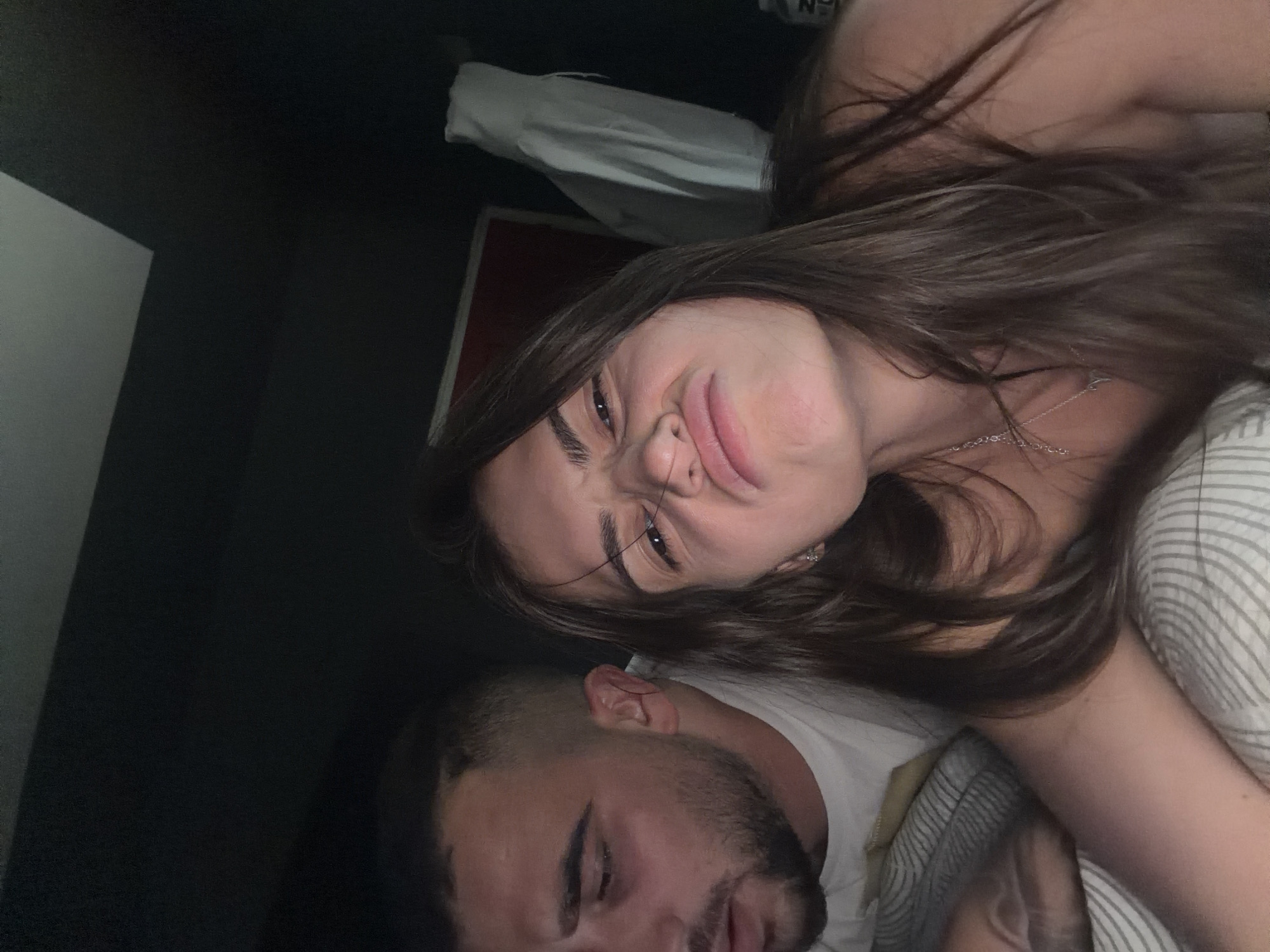 A close-up, indoor shot shows two individuals lying on a bed. A woman with long brown hair and a silver necklace is positioned towards the upper right. She is making a facial expression with her eyes squinted and nose wrinkled. A man with dark hair and a beard lies adjacent to her on the left side of the frame, with his eyes closed. They are on a bed with striped bedding. A white pillow and what appears to be a red object are visible in the background, along with a dark-colored wall. The location is Unknown, Unknown.