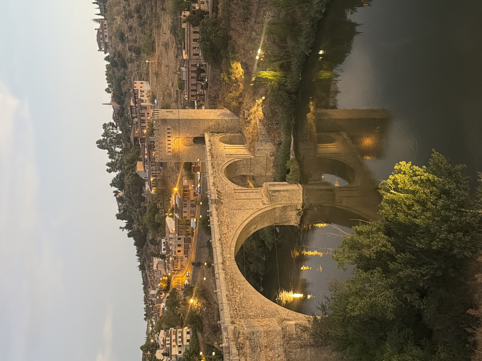 The image shows a view of the Puente de San Martín bridge in Toledo, Spain at dusk. The stone bridge, with its characteristic arches, spans across the Tagus River. A fortified tower is visible on the right side of the bridge. The river below reflects the lights from the bridge and the city. On the hillside overlooking the bridge, there are numerous buildings with illuminated windows, suggesting a residential area. Trees and vegetation are present on the riverbanks and the hillside. The sky is a soft twilight hue.
