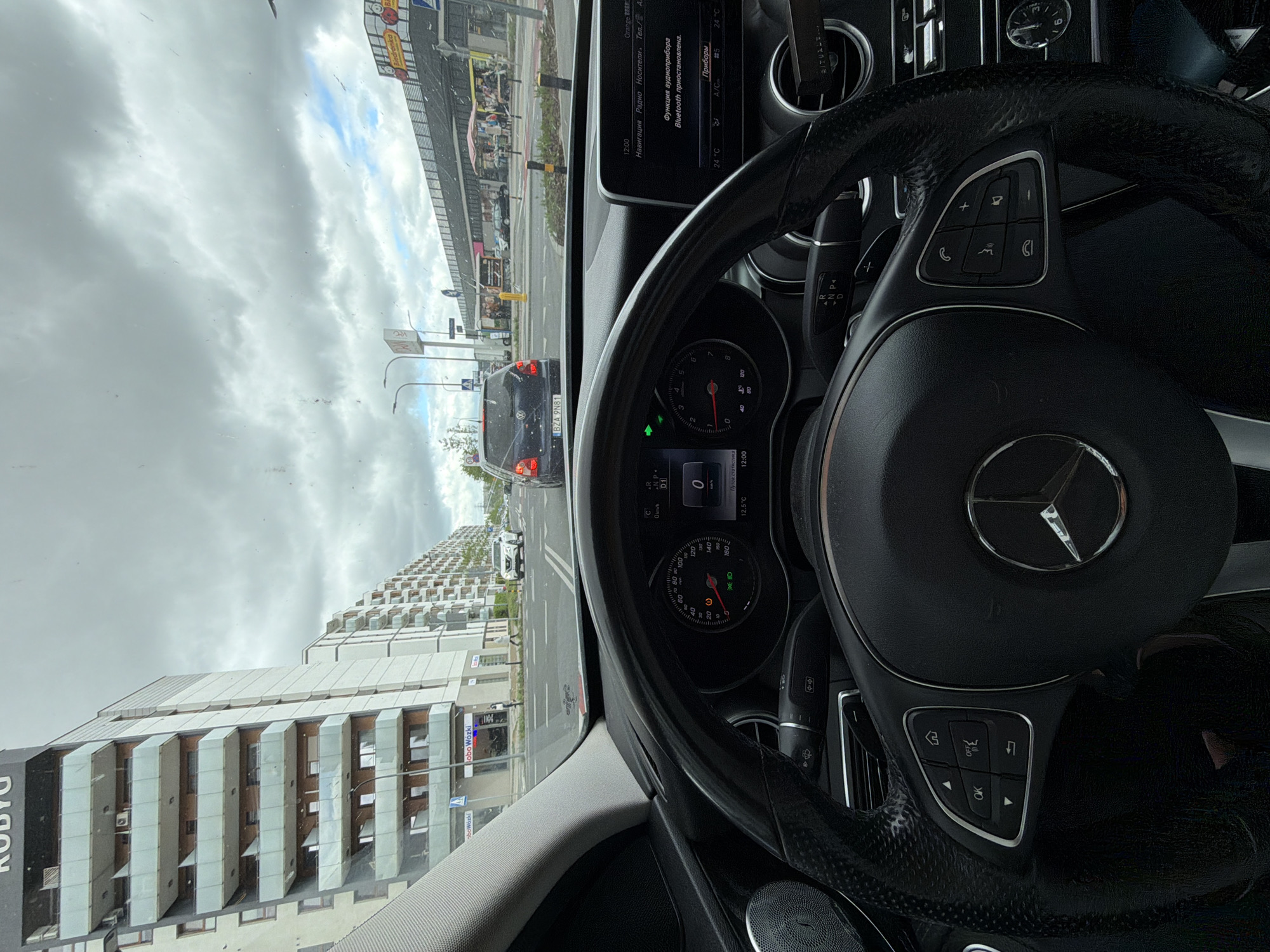 The image, taken from inside a car, shows an urban environment. The car is a Mercedes, as indicated by the steering wheel and dashboard. The speedometer shows 0 km/h, indicating the car is stationary. The license plate "BZA 9N81" is visible on the car directly ahead.
The environment includes a multistory building with "KO BYG" printed on its side. There is a sign "Bobo Wózki" visible, along with a pedestrian crossing. The sky is overcast. The car radio display indicates that the language is Russian and the temperature is 24°C or 12.5°C.