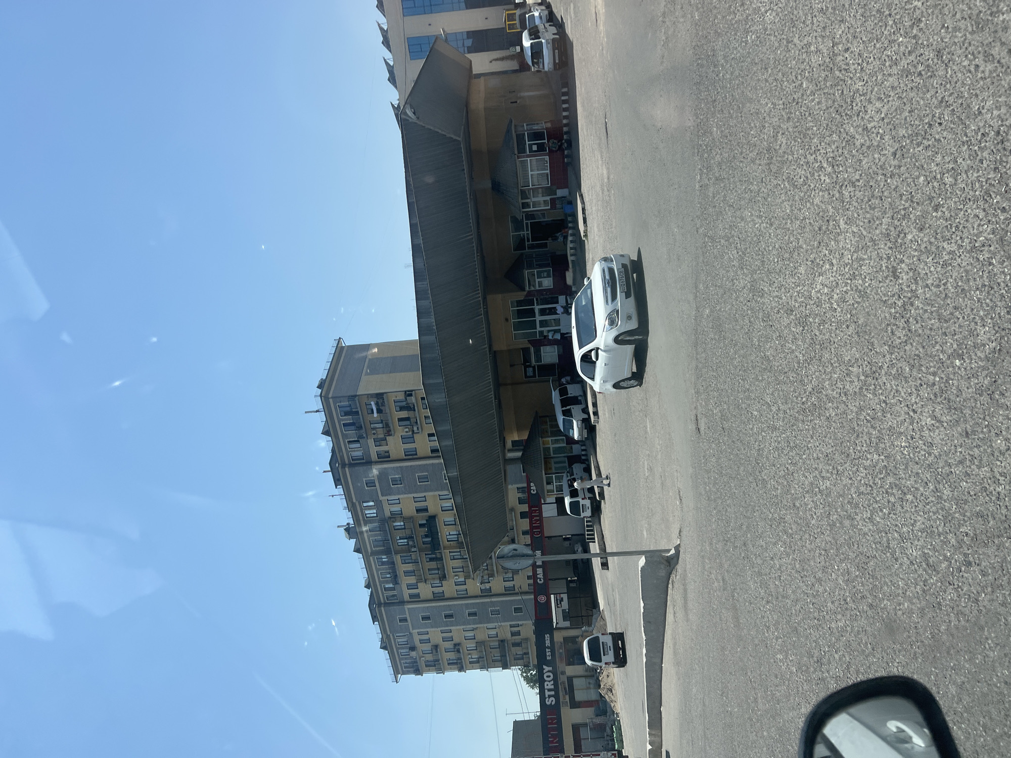 The image depicts a street scene in Kibray, Uzbekistan, on a clear, sunny day. In the foreground, a grey asphalt road is visible, with a portion of a car's side mirror on the left. A white sedan is driving on the road. Behind the road, a low-slung, covered market or commercial area with a distinctive slanted roof extends across the midground. Several people are seen walking near and inside the market area. A multi-story apartment building with a light-colored facade and balconies stands prominently in the background, to the left of the market. Several white vans are parked near the market. The overall environment suggests a commercial district in a town or city.